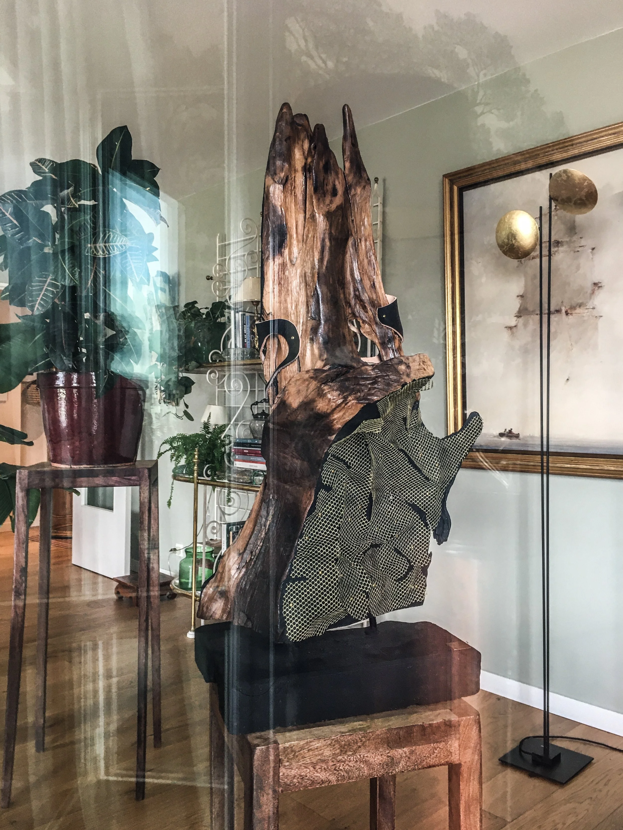 Une sculpture en bois avec des éléments métalliques, placée dans une pièce avec des plantes et des œuvres d'art, visible à travers une fenêtre ou une vitre.