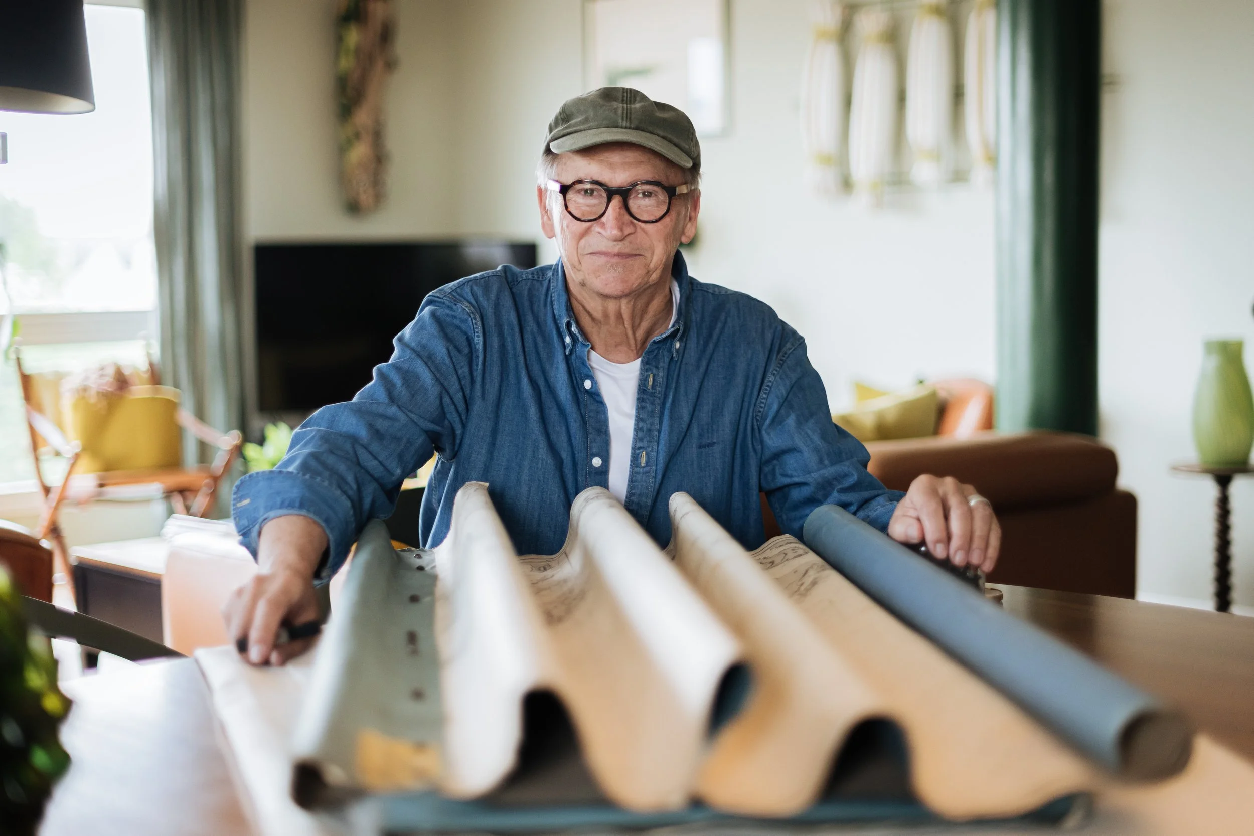 Un homme âgé avec lunettes, portant une casquette et une chemise en denim, assis à une table devant un rouleau de papier.
