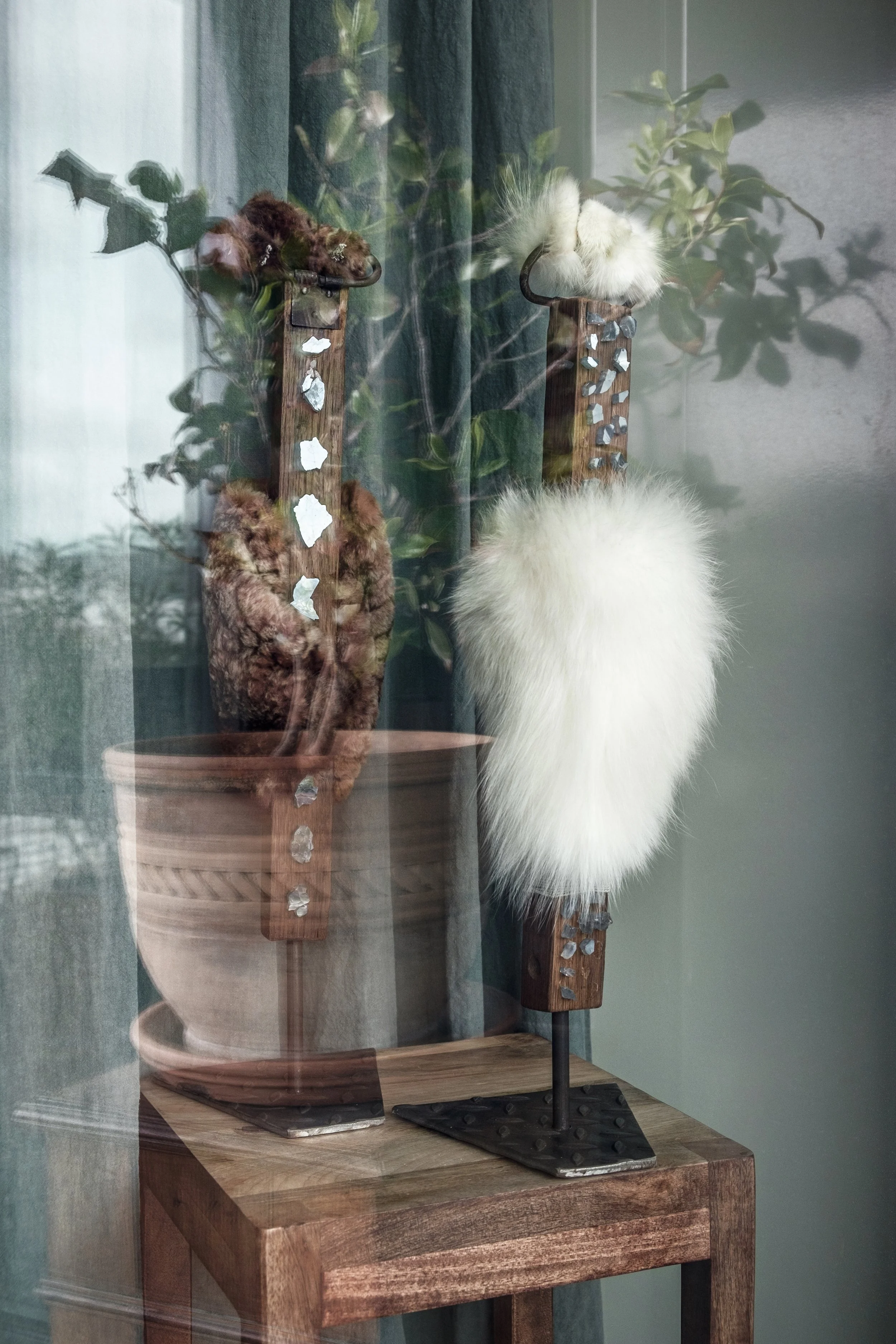 Deux tapis résille en bois avec fourrures, l'une brune et l'autre blanche, exposés dans une vitrine avec des plantes en arrière-plan.
