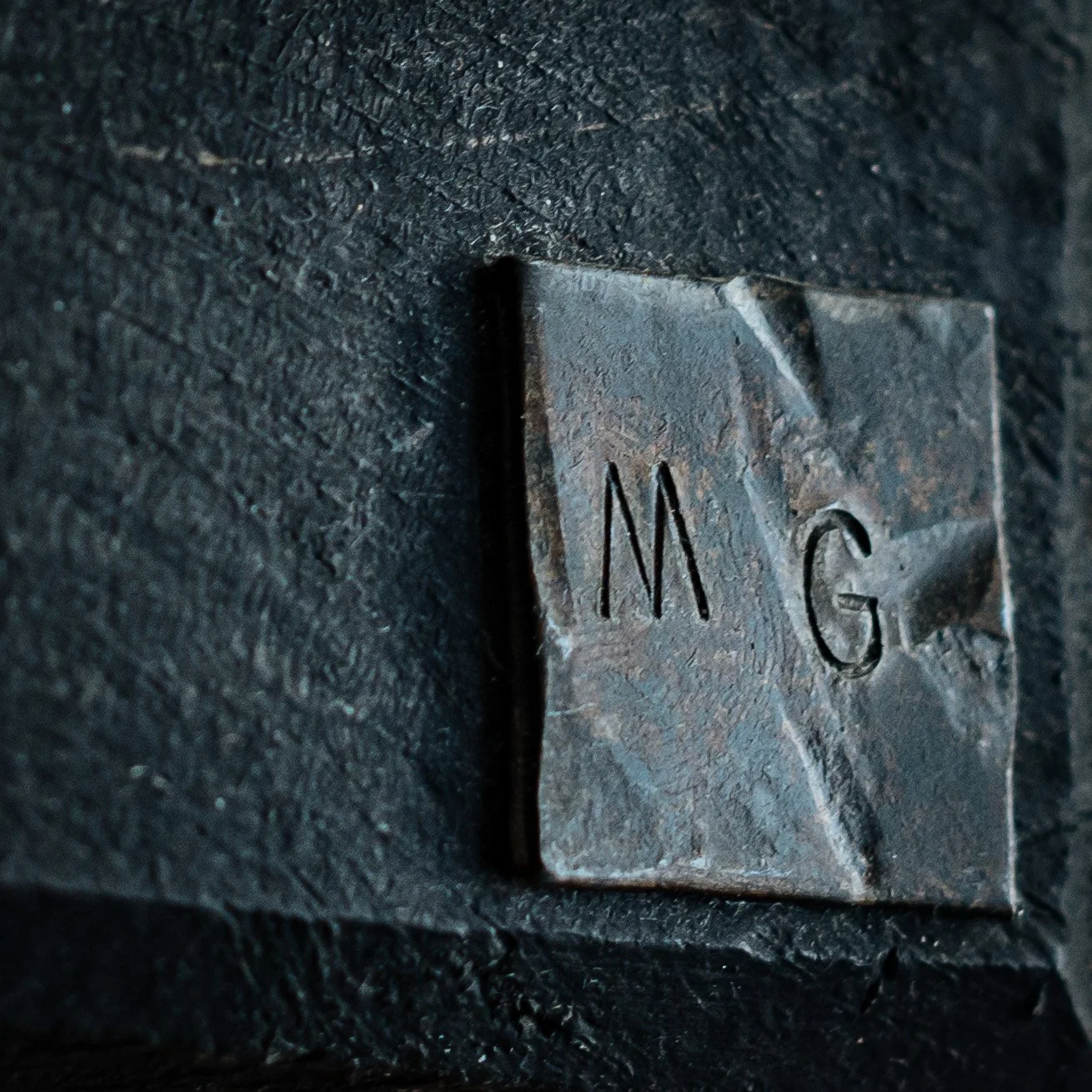 Une plaque en métal rouillée avec les lettres « M G » gravées, fixée sur une surface sombre en bois.