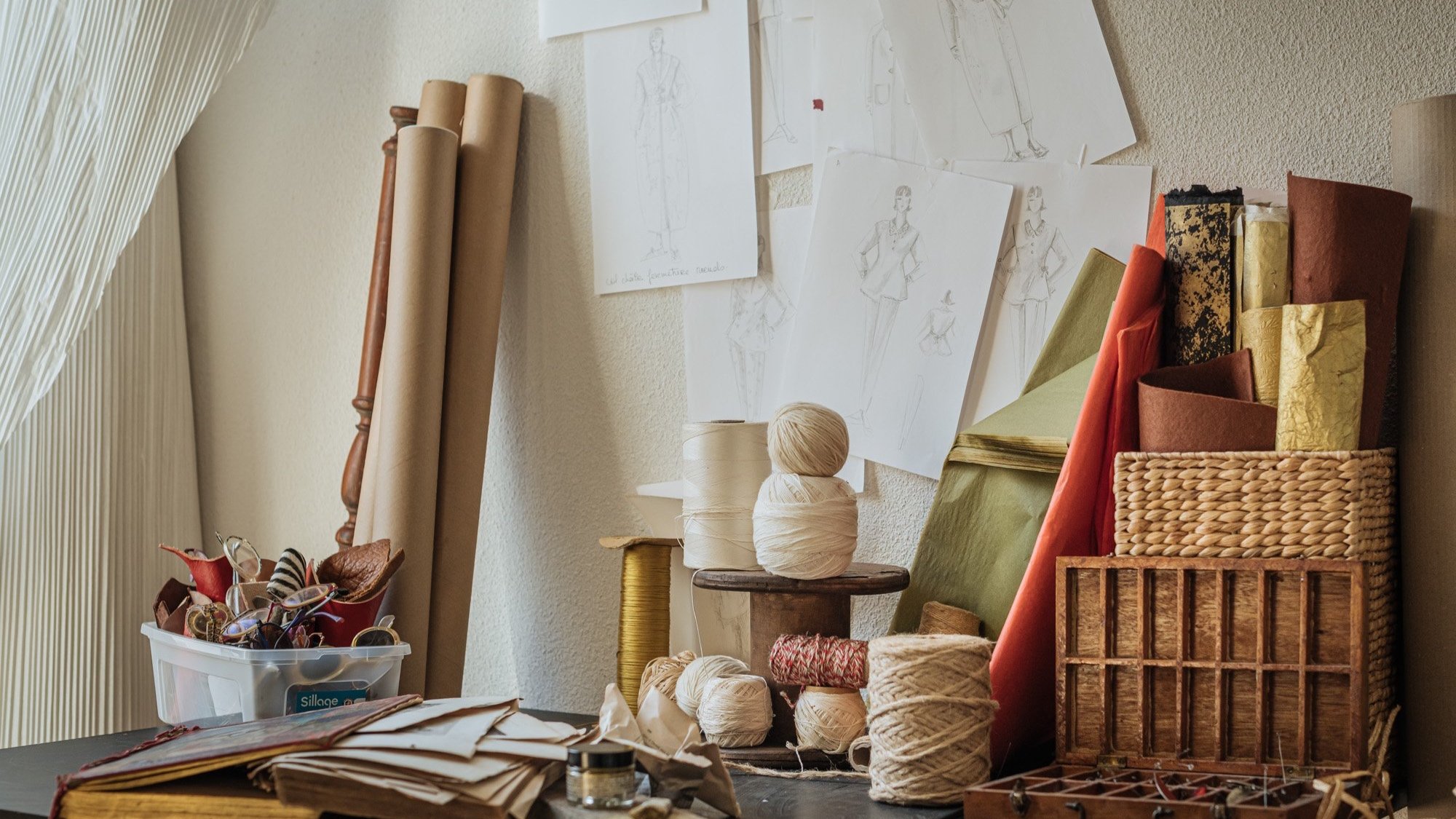 Espace de travail de couture avec échantillons de tissus, pelotes de fil, croquis de mode sur le mur, et divers outils de couture.