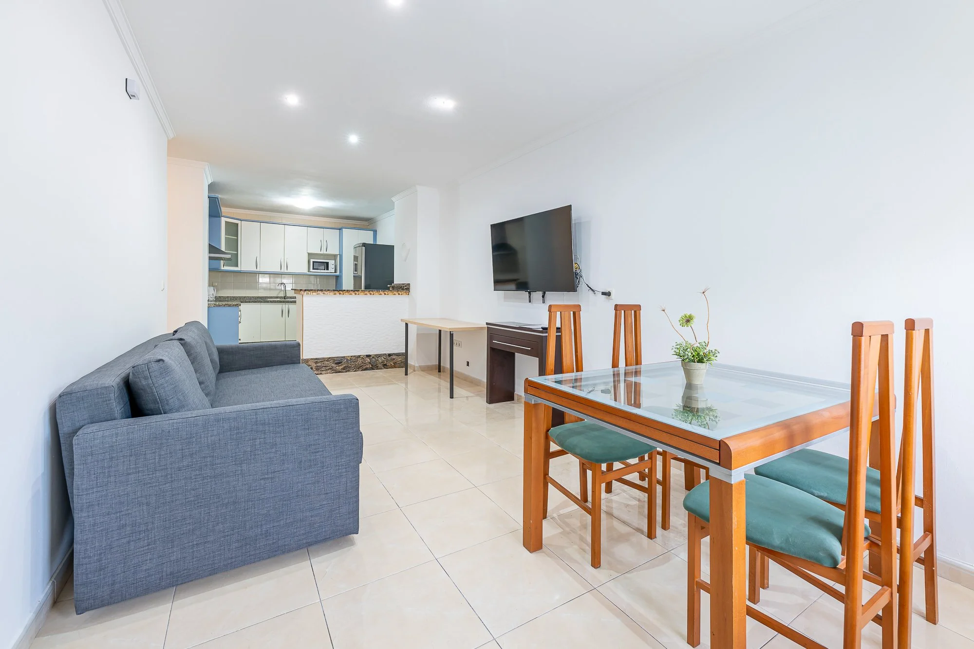 SpainBreaks | Living room and kitchen area with gray sofa-bed (sleeps 2), glass dining table with four wooden chairs, a remote work desk with 3 electrical sockets, wall-mounted 55 inch smart TV.