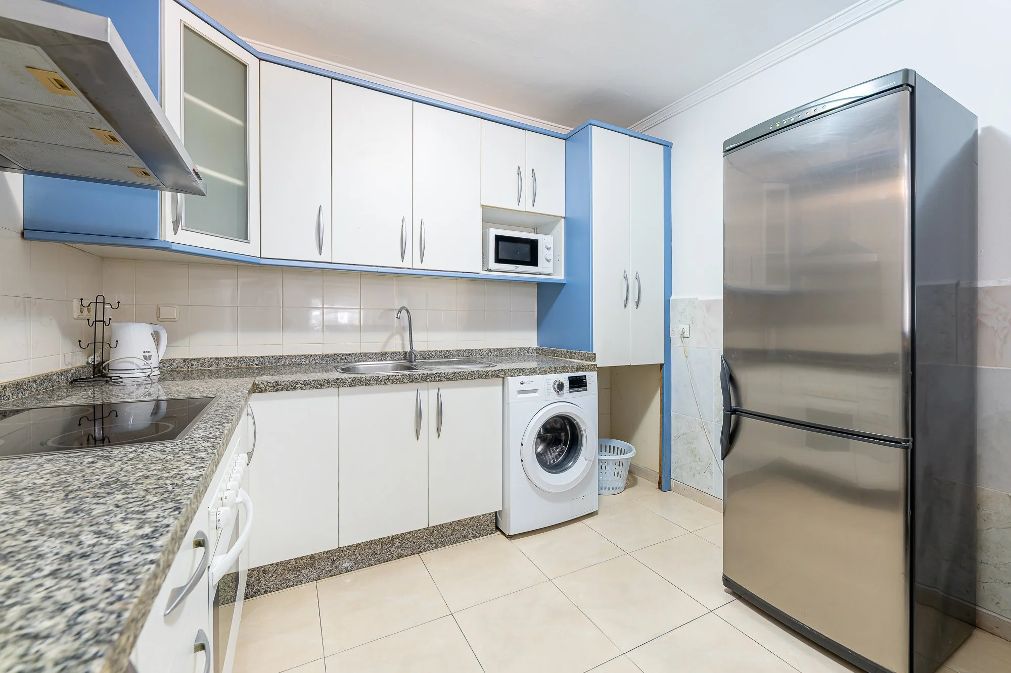 SpainBreaks | Kitchen with granite countertops, white and blue cabinets, a microwave, washing machine, refrigerator, and small appliances.
