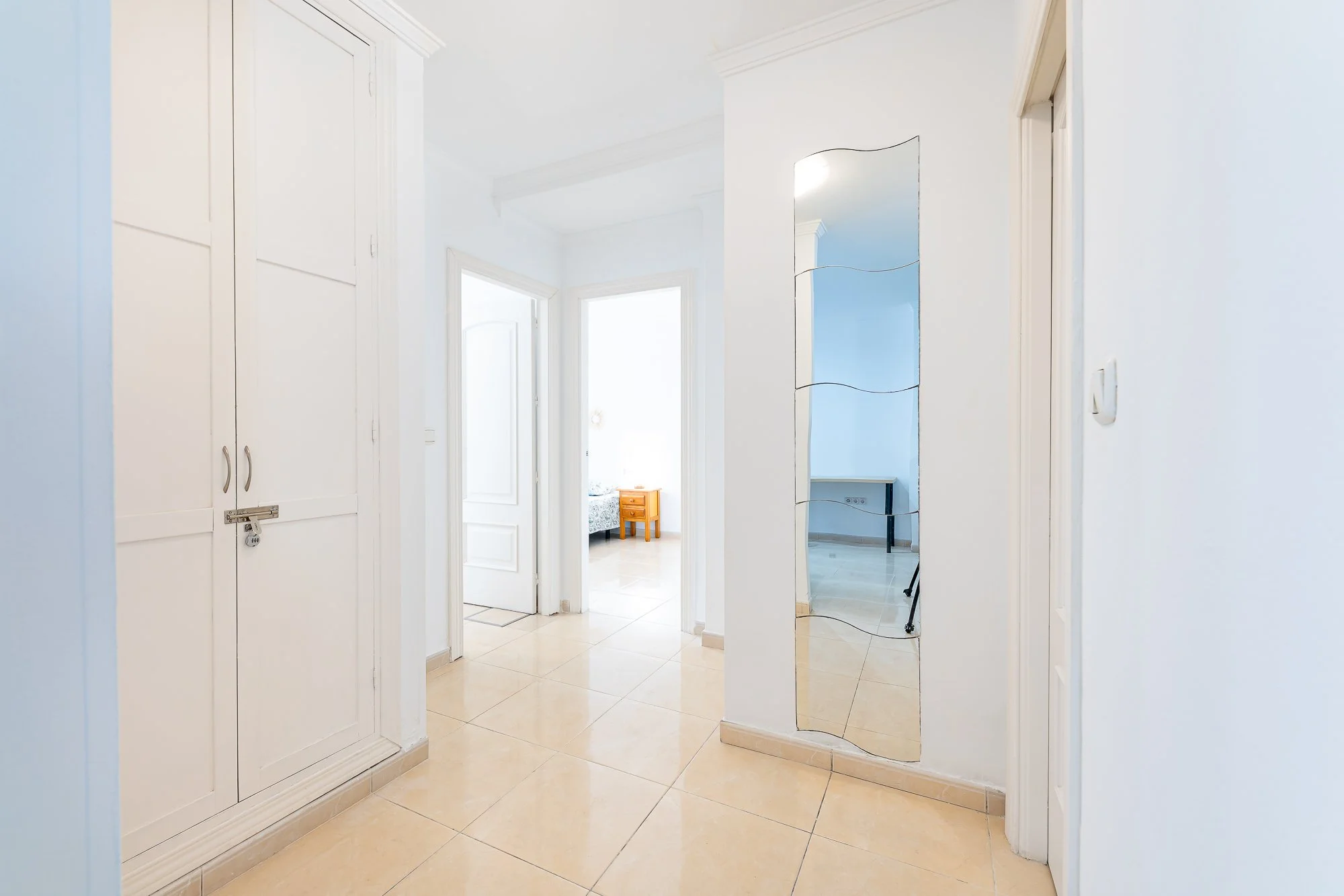 SpainBreaks | Bright hallway with white walls, beige tile floor, curved mirror on the right, white wardrobes on the left and doors leading to the bedrooms and bathroom.