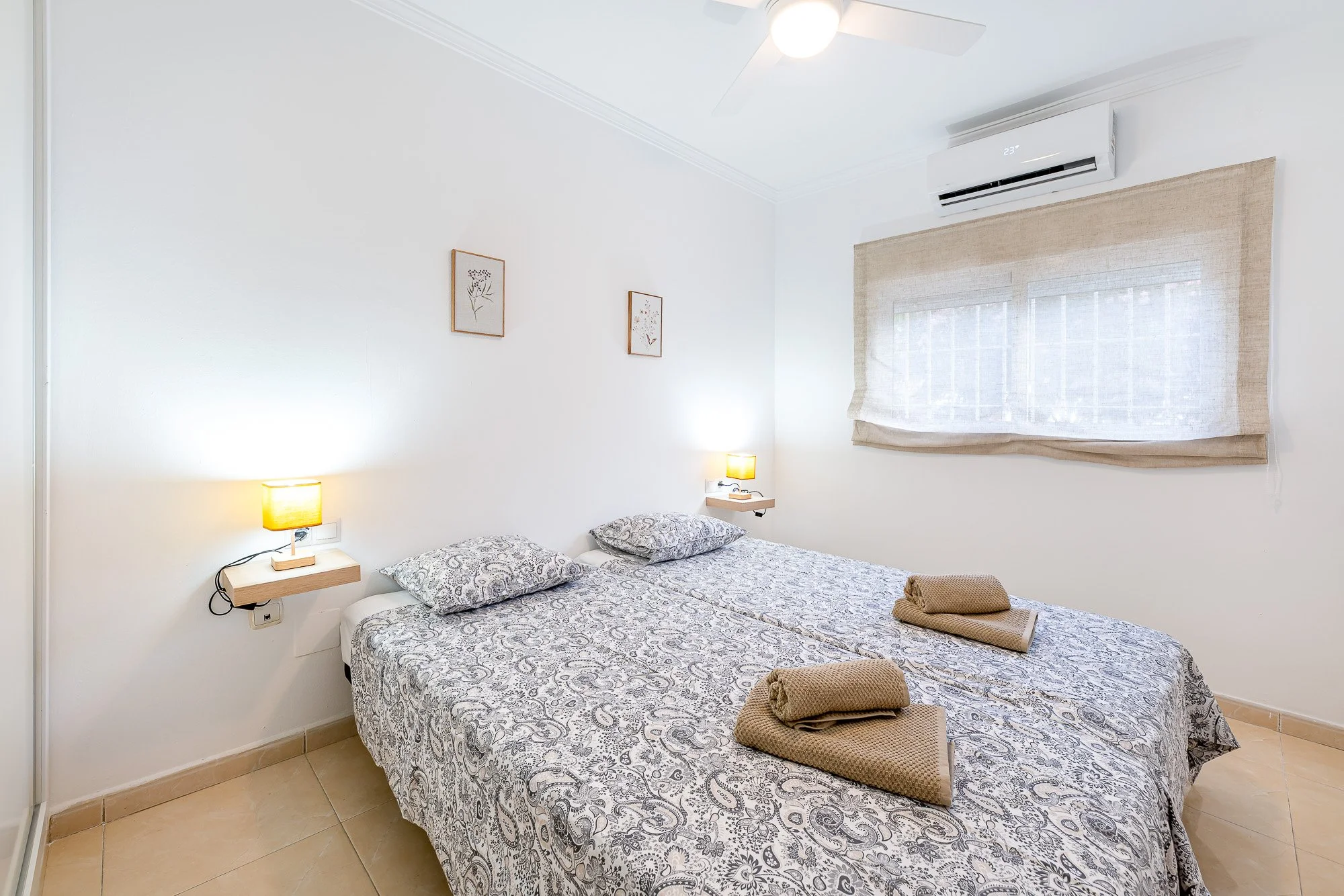 SpainBreaks | Bedroom 2 - white walls, and a split air conditioning unit. There are two single beds pushed together. Wall-mounted nightstands with lamps beside each bed and USB sockets.