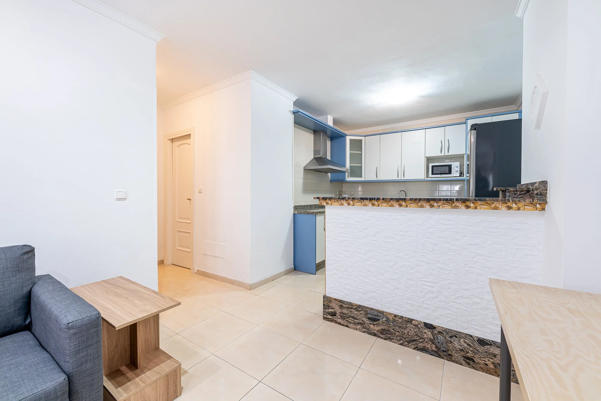 SpainBreaks | Living room and kitchen in a modern apartment with white walls, beige floor tiles, gray sofa-bed (sleeps 2), wooden side table, and a partially open kitchen with white cabinets, granite countertops, a microwave, oven, cooker, washing ma
