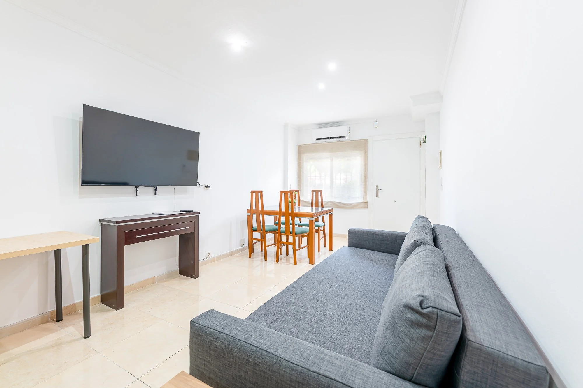 SpainBreaks | Living room with work-from-home desk, gray sofa-bed (sleeps 2), wall-mounted flat-screen 55 inch smart TV, a dining table with four chairs, air conditioning unit, and white walls.
