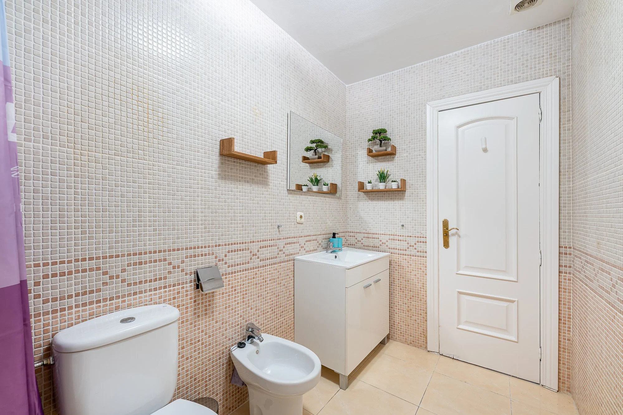 SpainBreaks | Bathroom with beige tiled walls, toilet, bidet, sink cabinet, mirror and wooden shelves.