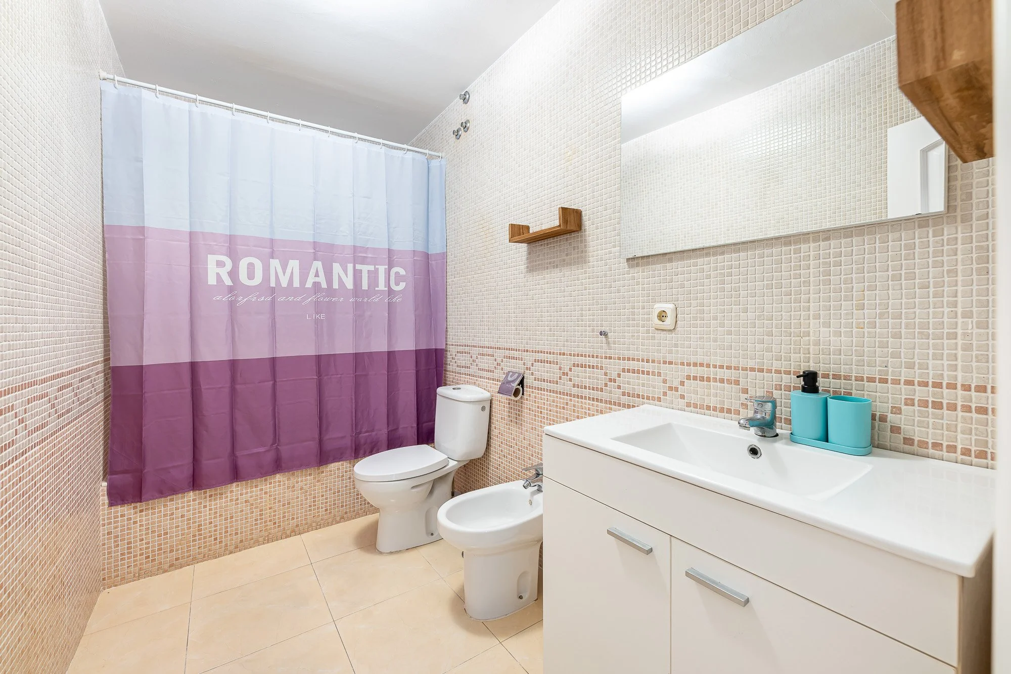 SpainBreaks | Bathroom with a shower curtain, toilet, bidet, sink with a mirror, and wooden shelves.