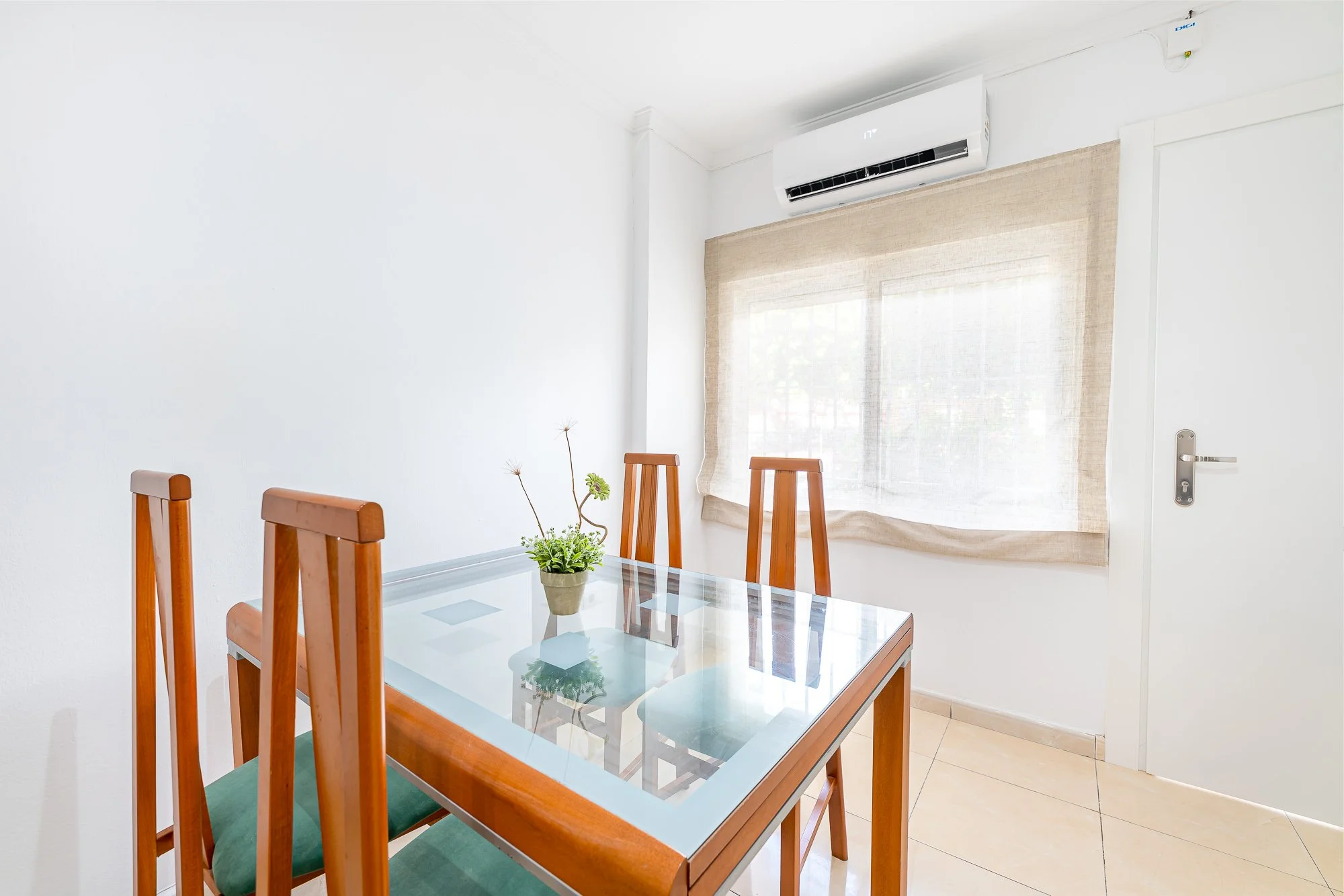 SpainBreaks | Dining room with four wooden chairs around a glass-top table, and an air conditioning unit above the window.