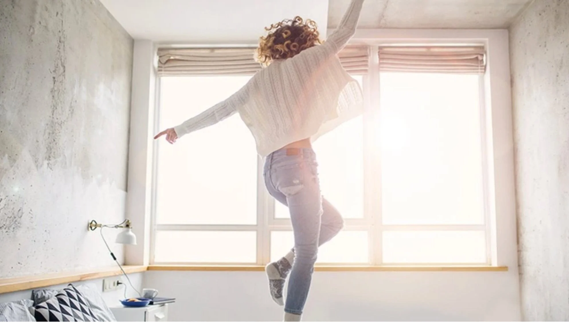 Person jumping on bed in a brightly lit room with large windows and minimalist decor.