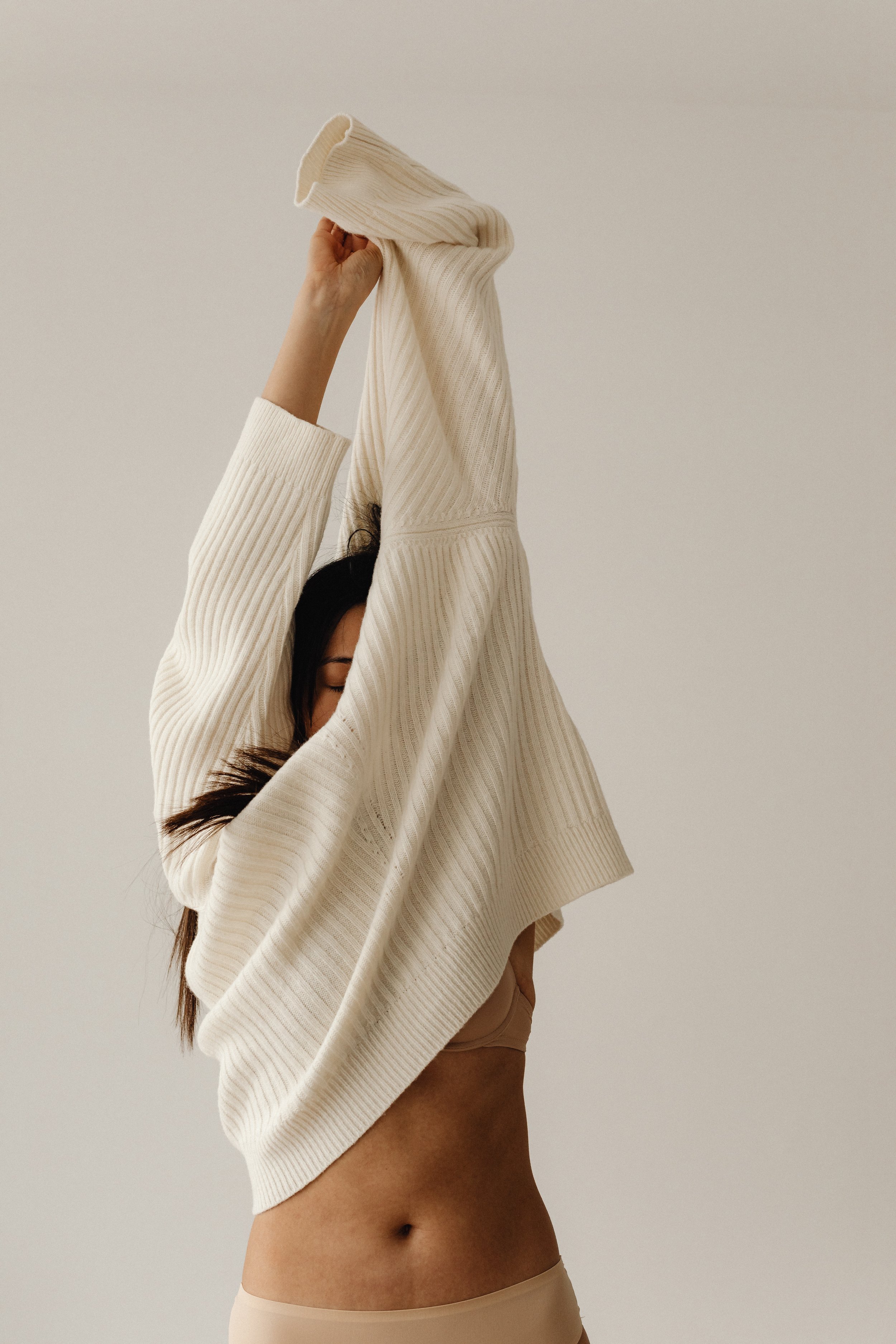 Femme levant un pull en laine beige, habillée d'un sous-vêtement beige, dans un décor neutre.