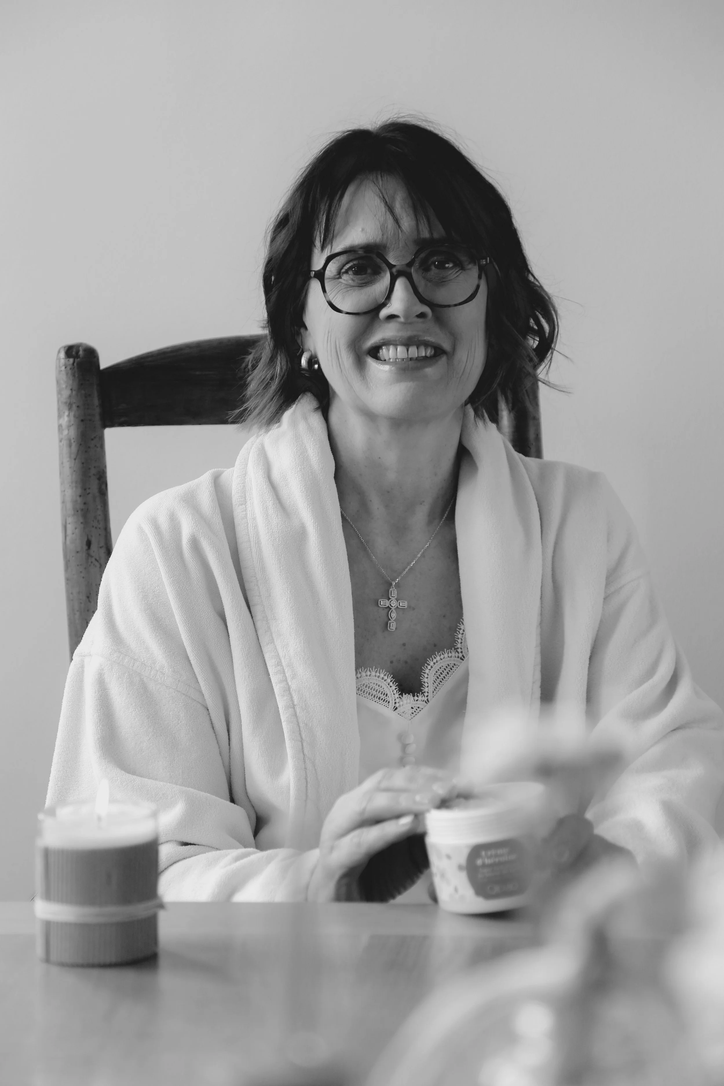 Photo en noir et blanc d'une femme assise à une table, portant des lunettes et un châle, tenant un pot de yaourt avec une cuillère, avec une bougie allumée sur la table.