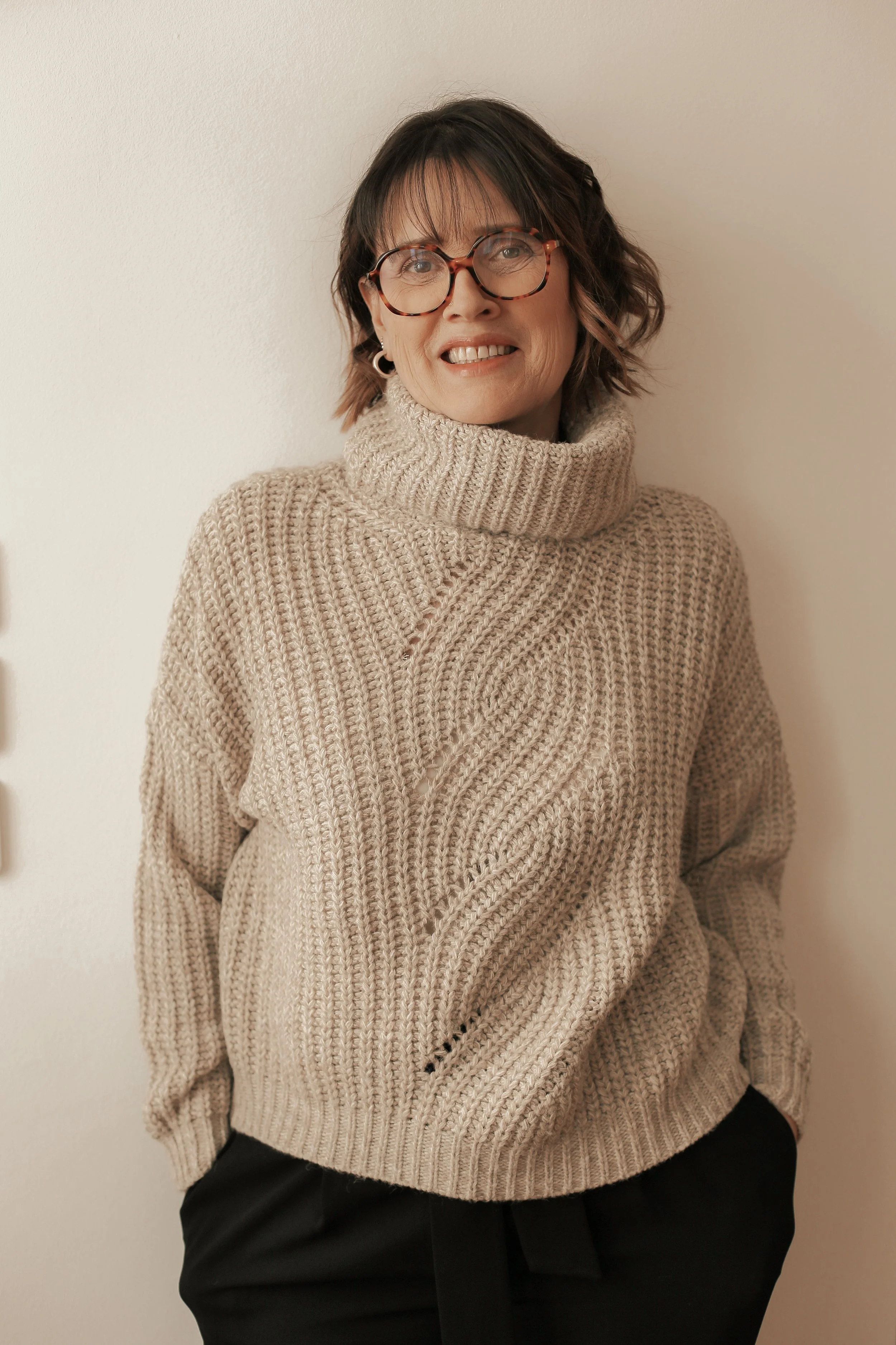 Femme portant un pull en maille beige avec un col roulé, cheveux bruns ondulés, portant des lunettes rondes, souriant, contre un mur blanc.