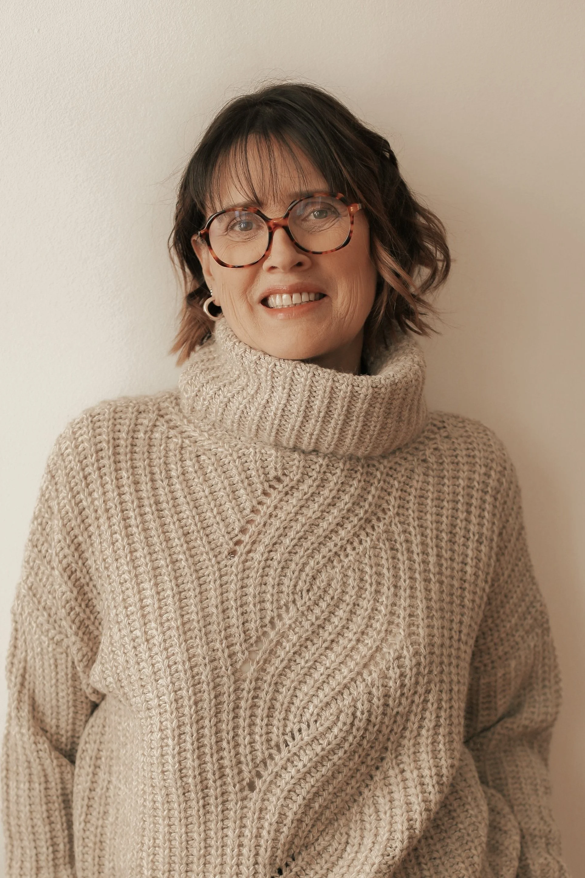 Une femme avec des lunettes rondes, portant un pull en laine beige à col roulé, souriant, devant un mur blanc.