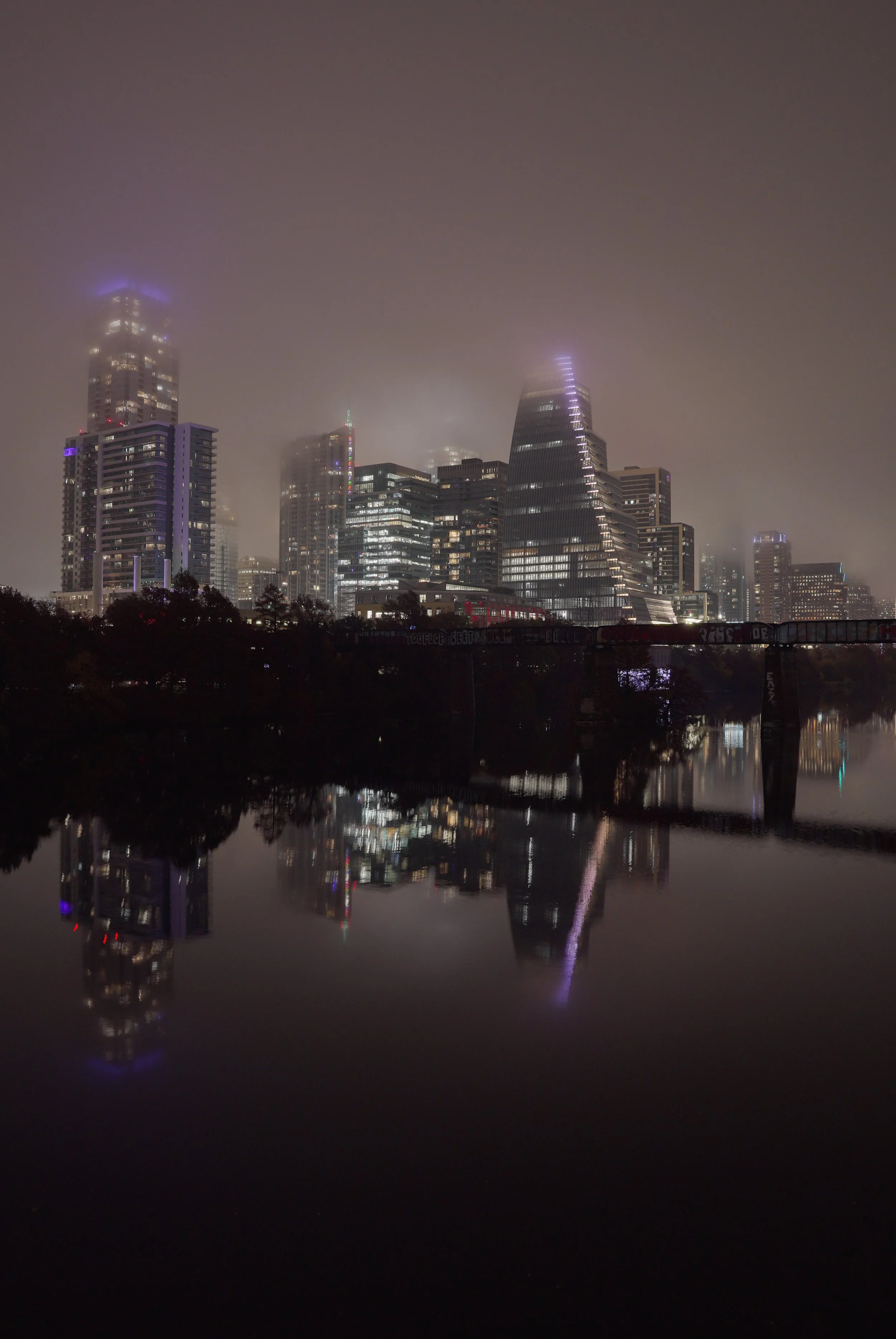 Nighttime cityscape of Austin Texas with illuminated skyscrapers partially obscured by fog, reflected in a calm body of water in the foreground.