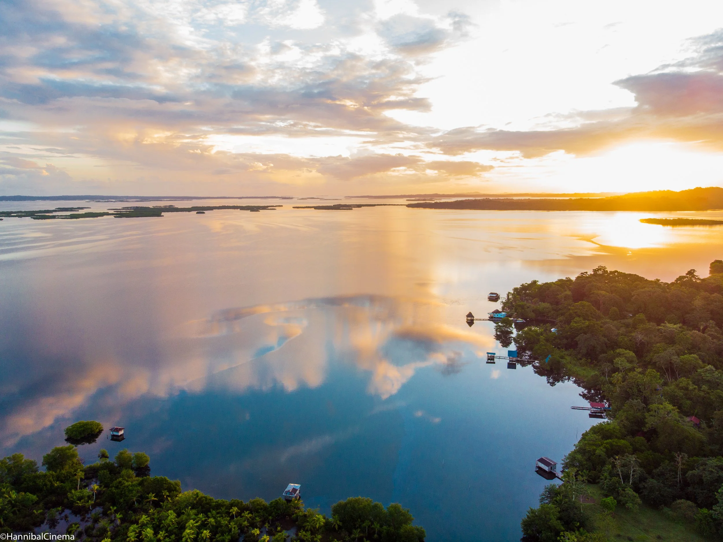 Aerial view of a river at sunset with reflections of clouds in the water, surrounded by lush green trees and small houses on the shoreline.