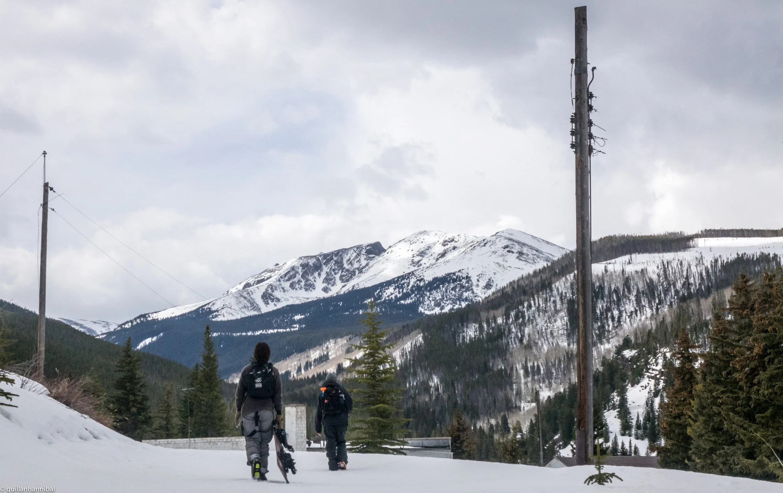 Two hikers walking on snow-covered ground through a mountain landscape with snowy peaks, pine trees, and an overcast sky in Colorado.