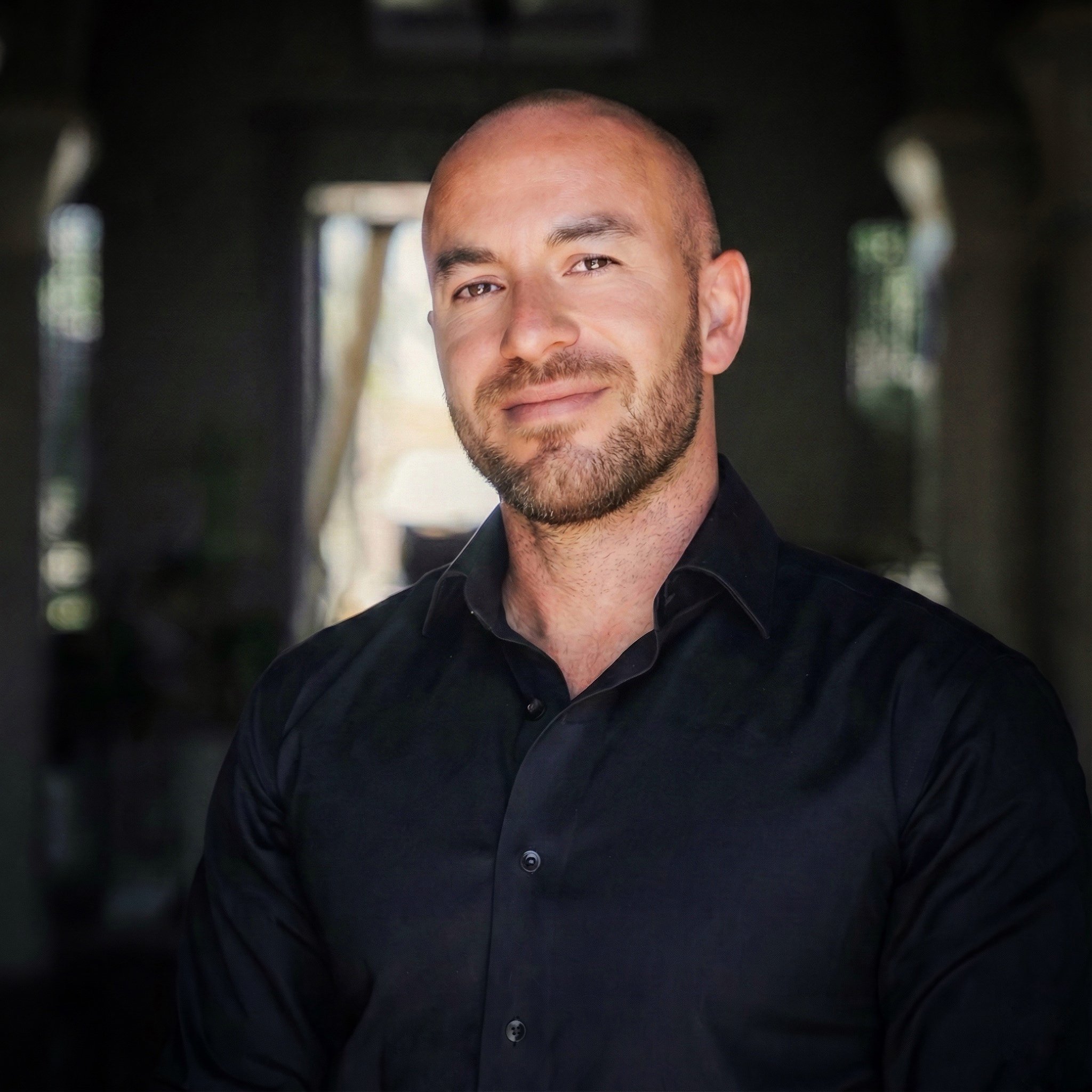 A man with a beard and short hair wearing a black shirt, smiling softly at the camera, standing in a dark, rustic setting with blurred background windows.