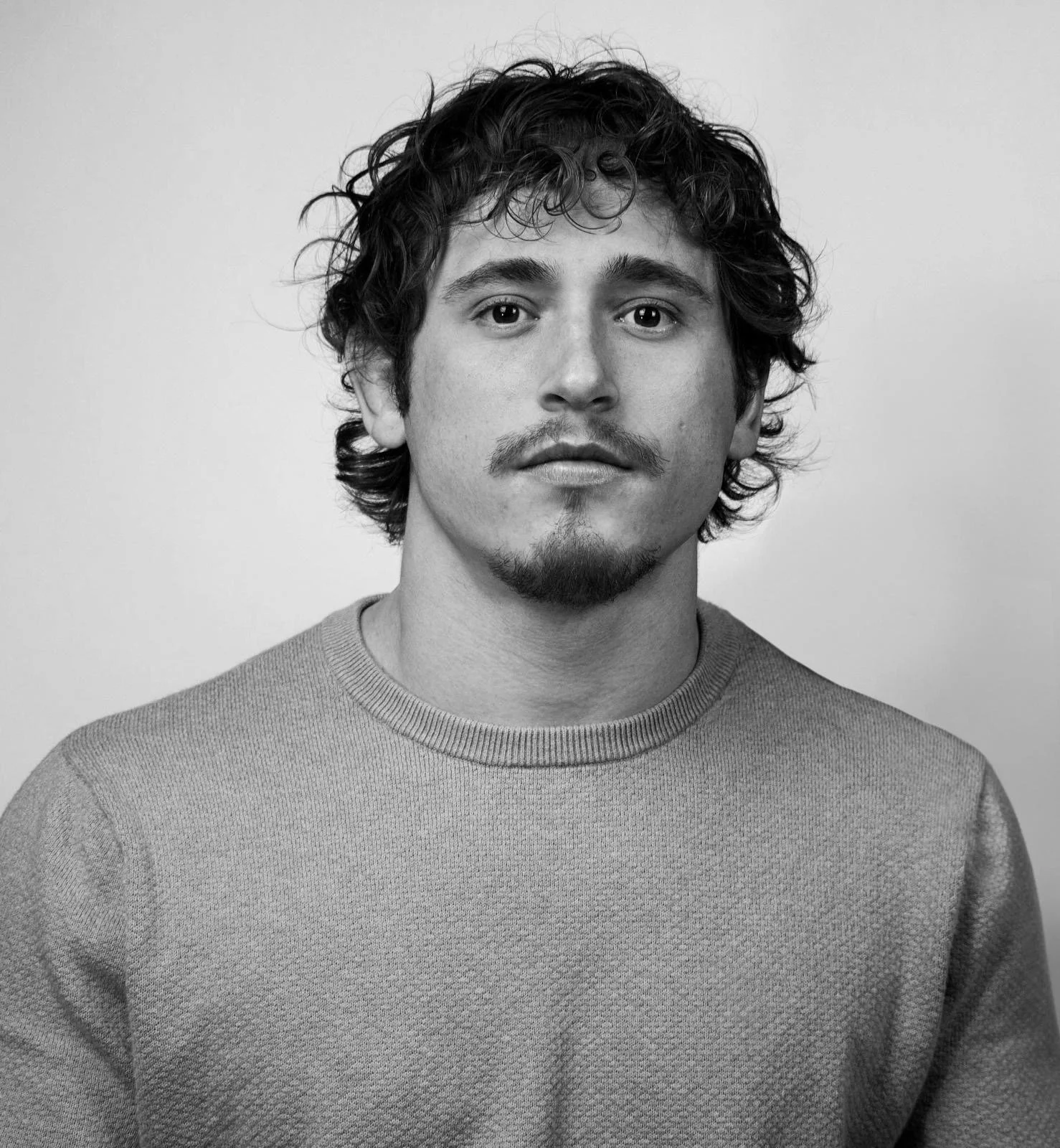 Black and white portrait of a young man with curly hair, a mustache, and a goatee, wearing a textured crew neck sweater, looking directly at the camera.