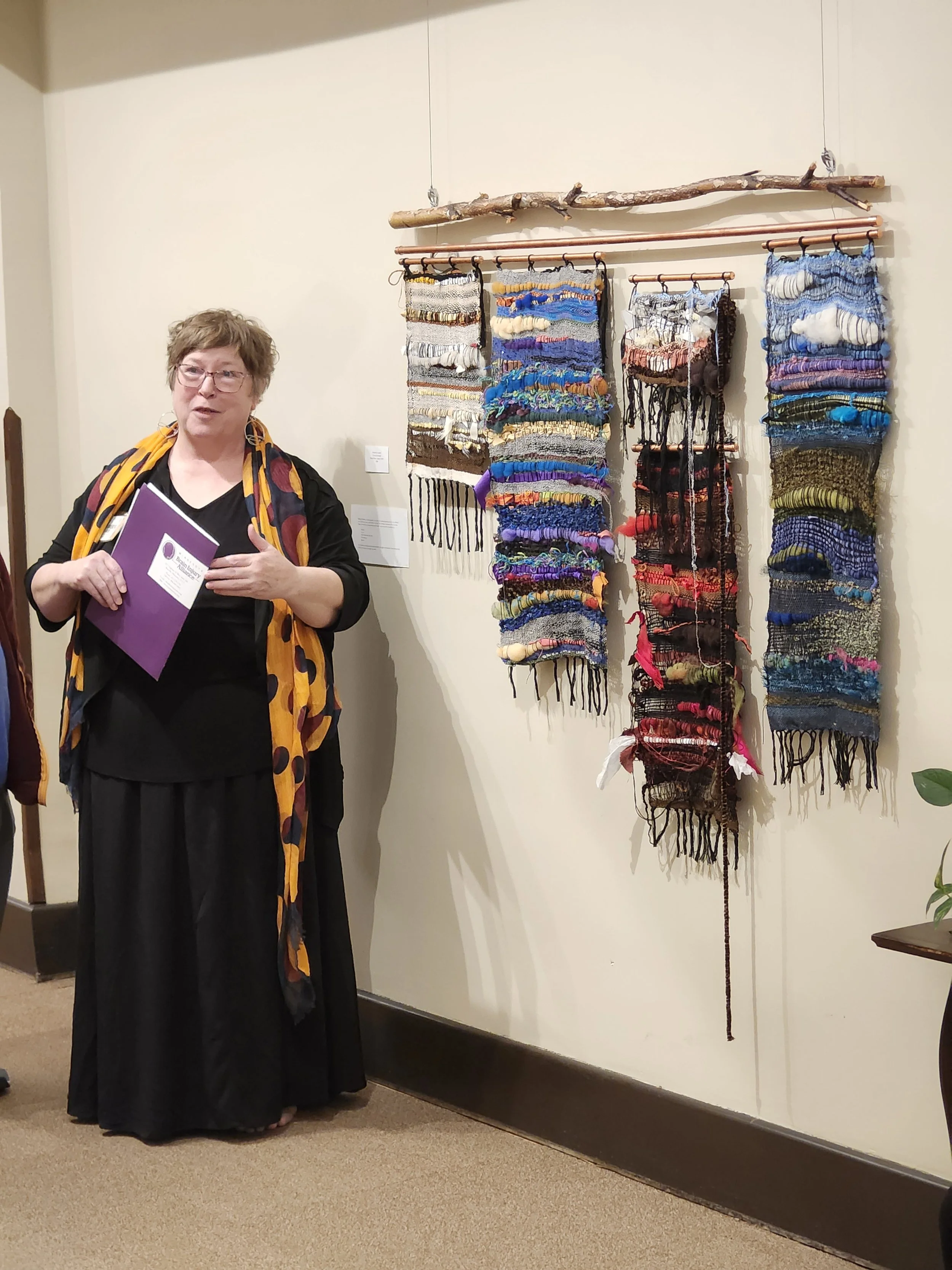 White woman with short brown hair in a black dress standing next to woven fiber wall art.