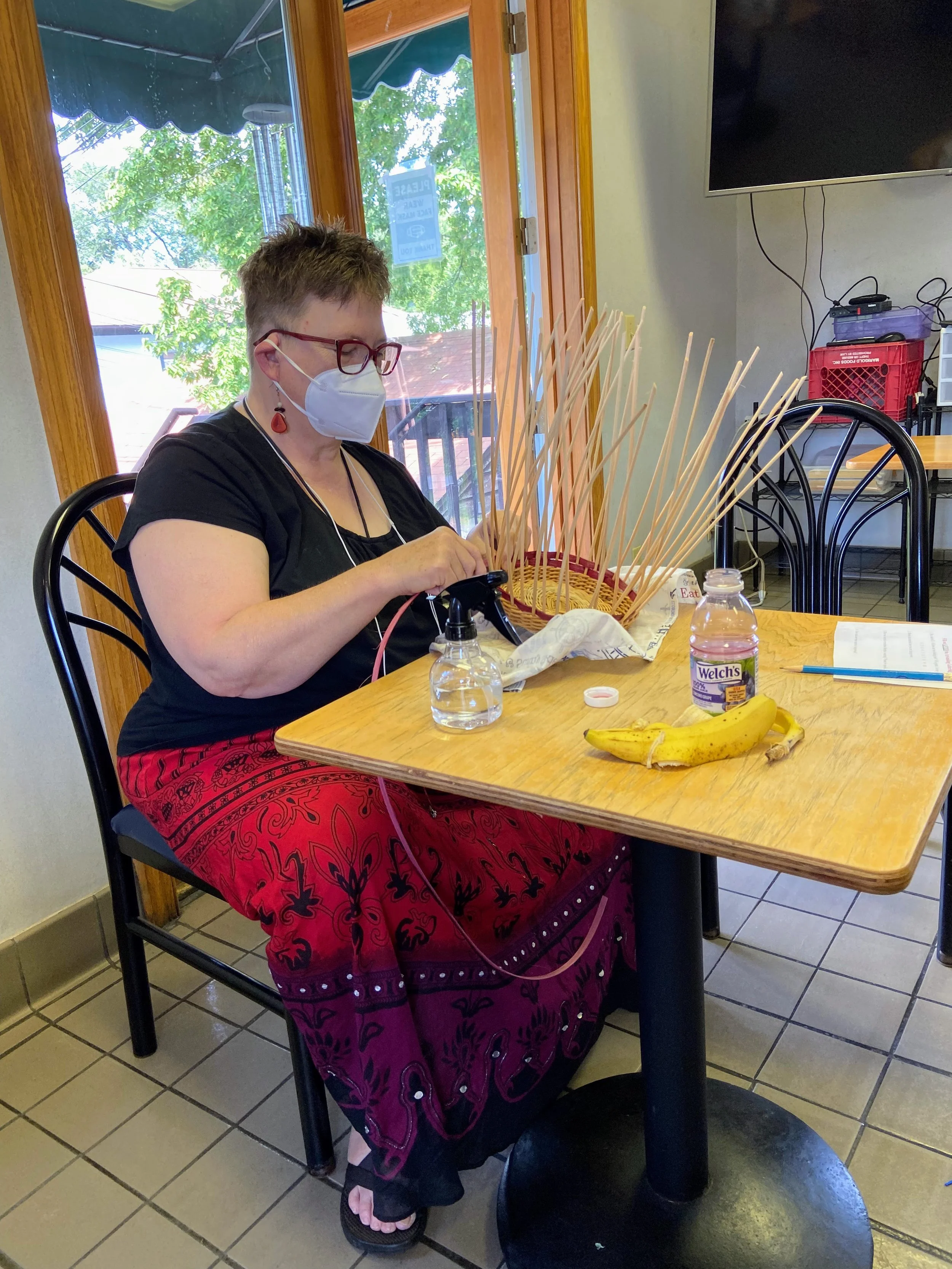 White woman with short hair in a red skirt sitting at a table weaving a basket.