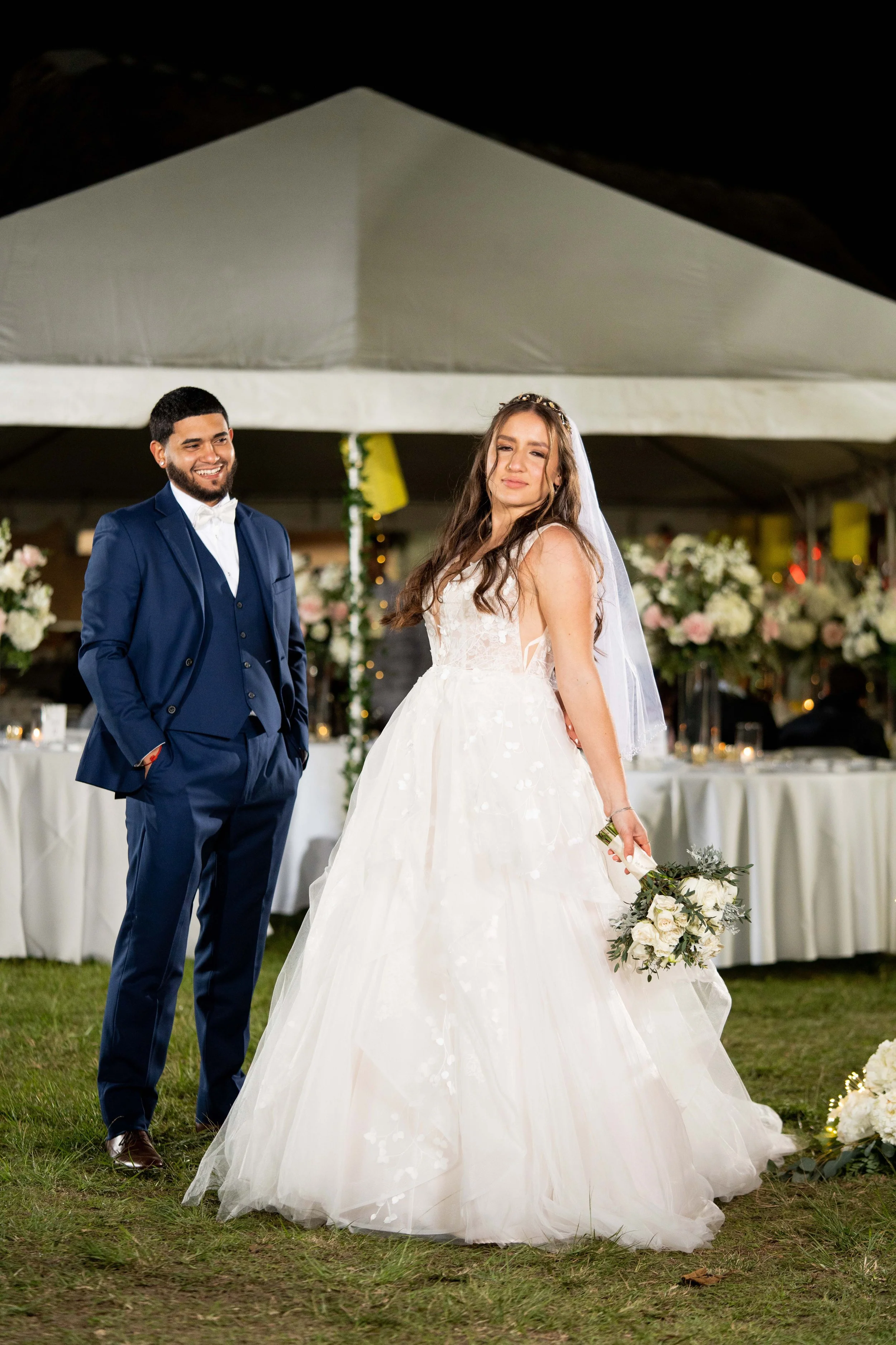 A bride and groom in wedding attire standing outdoors, with the bride holding a bouquet of white flowers. They are in front of a decorated tent with floral arrangements.