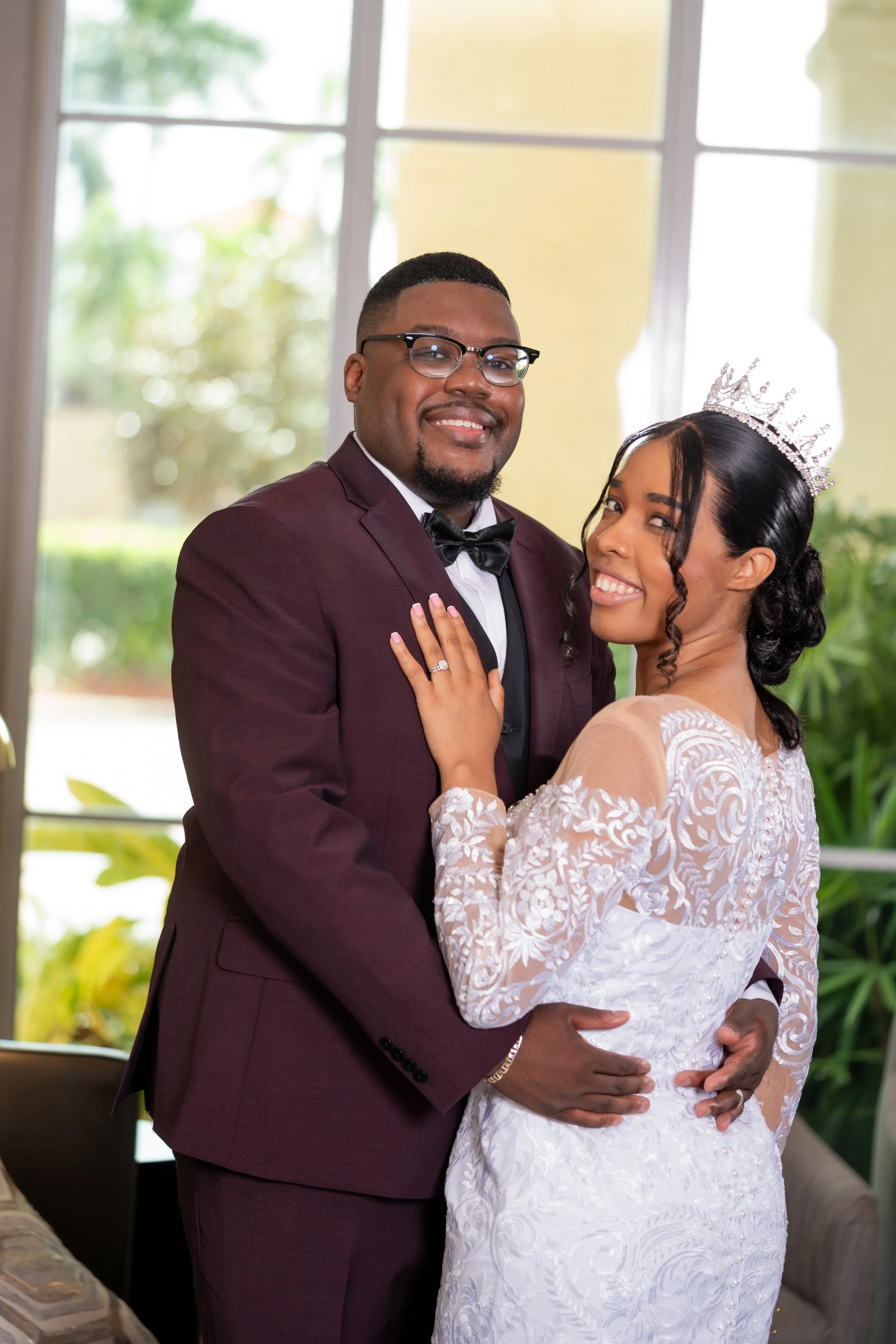 A bride and groom posing indoors near a window, with the bride wearing a white lace gown and tiara, and the groom in a burgundy suit.
