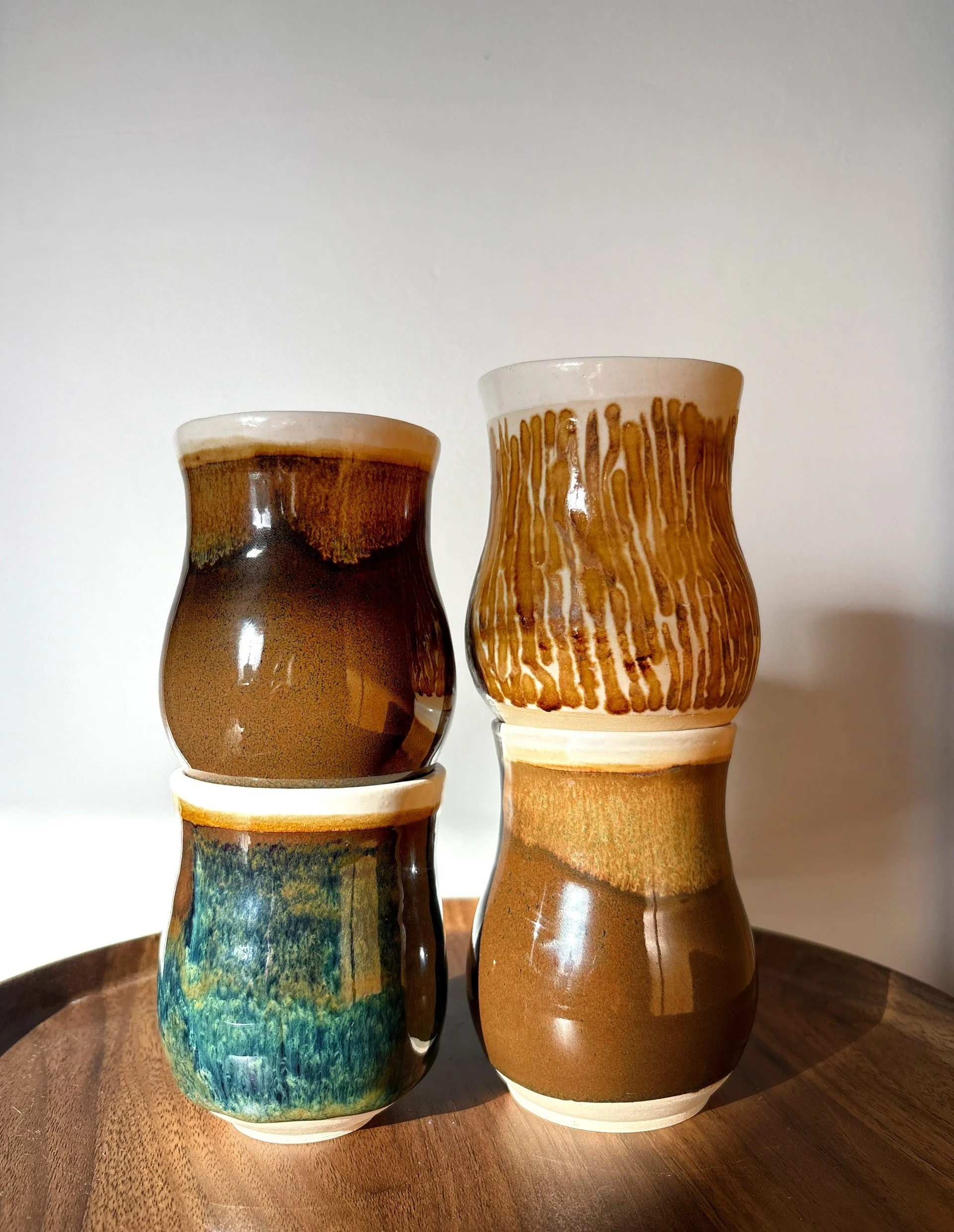 Four stacked ceramic vases with earth tone glazes on a wooden surface against a plain white wall.