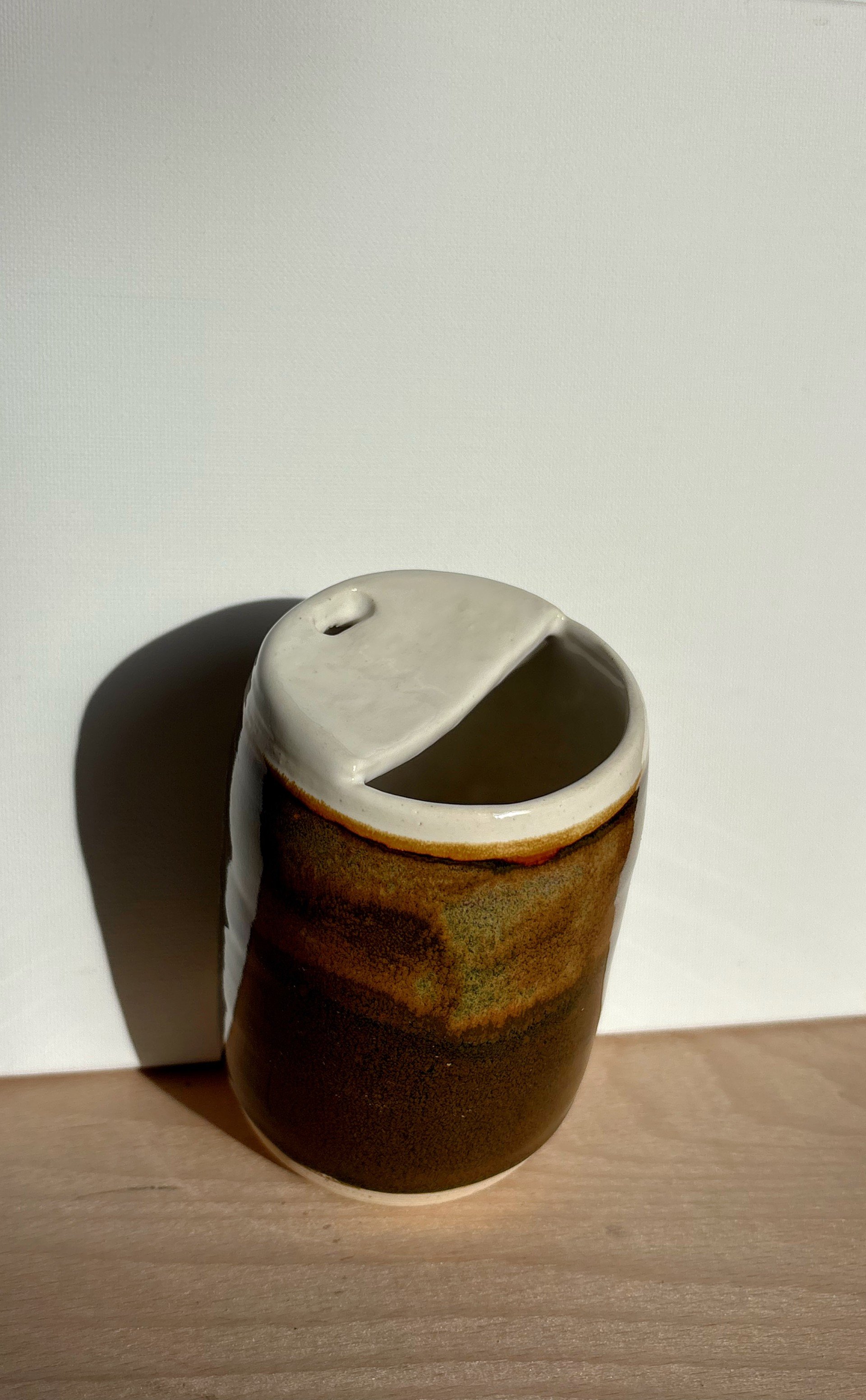 Ceramic travel cup with a white, half-open lid and rustic brown glaze, placed on a wooden surface against a plain light-colored background. Inspired by a coocnut glaze 