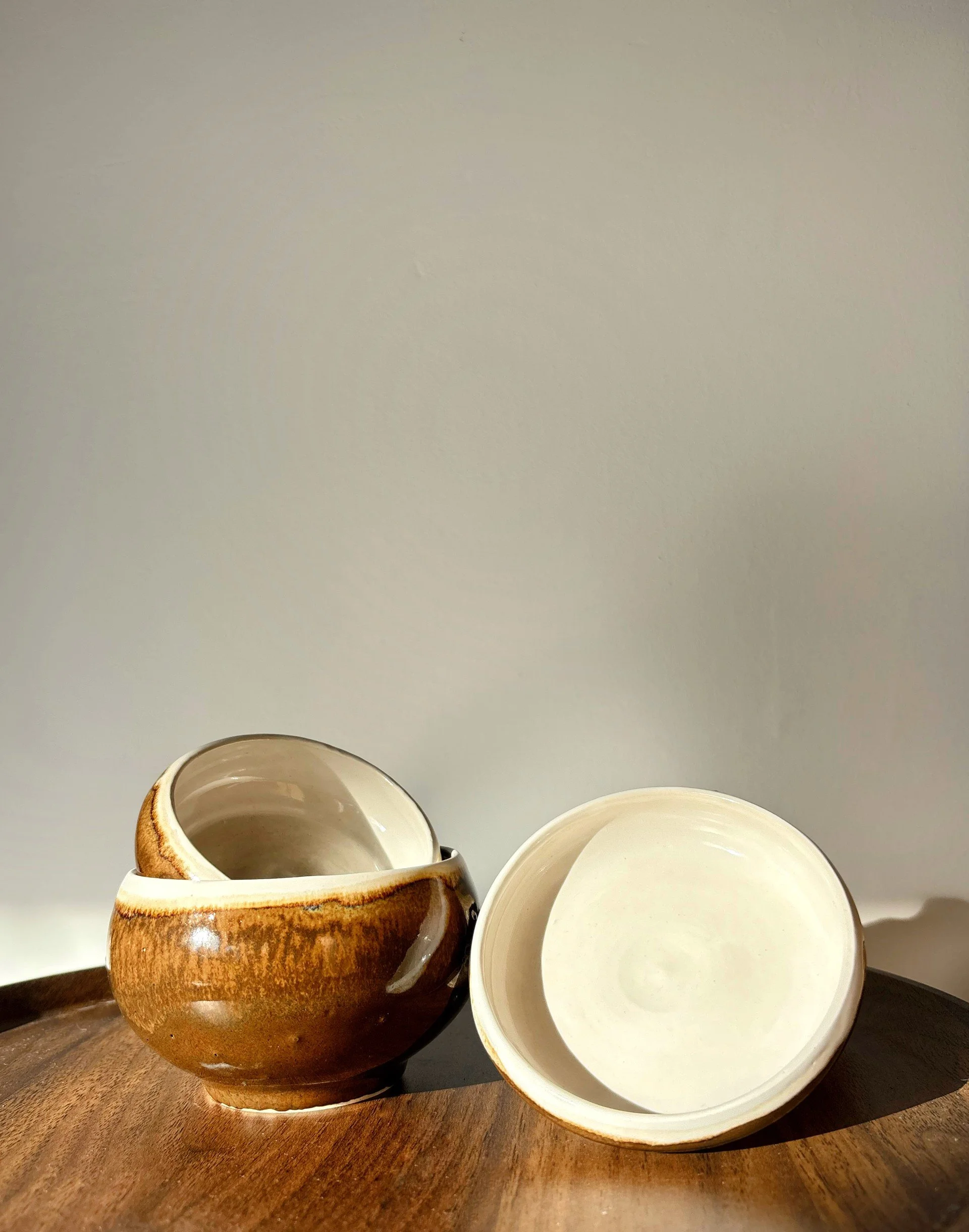Two ceramic bowls, one brown and one white, placed on a wooden surface against a plain wall. Inspired by coconuts with  a brown body and white top. Captures under the sunlight. They're unique, creative, aesthetic. 