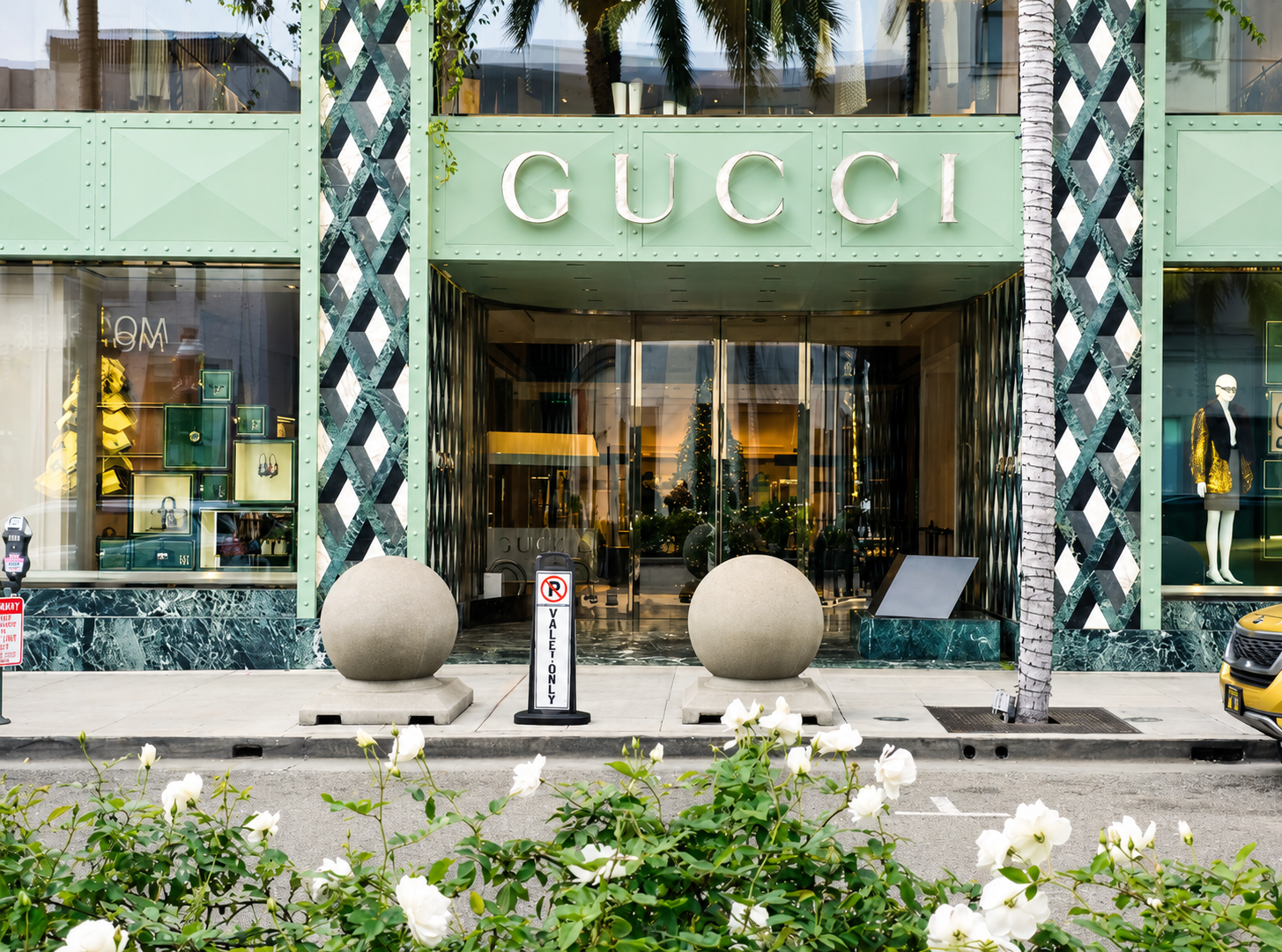 Facade of a Gucci store with large glass windows, green exterior, and prominent Gucci sign above the entrance. Two large spherical planters outside, a tree to the right, and white flowers in the foreground, with a parking sign and a yellow car partially visible.