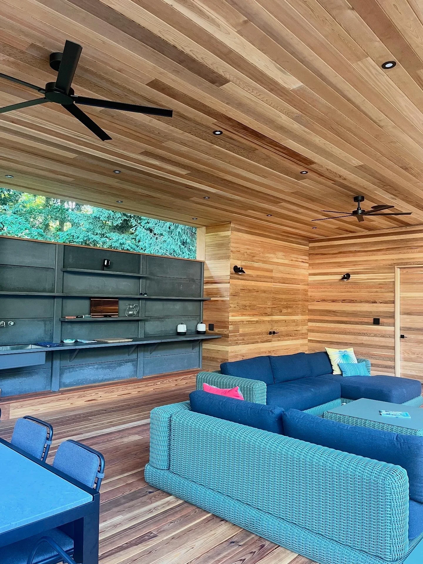 Interior of a room with wooden walls and ceiling, ceiling fans, a blue sofa, a wicker chair, and a large window showing trees outside.