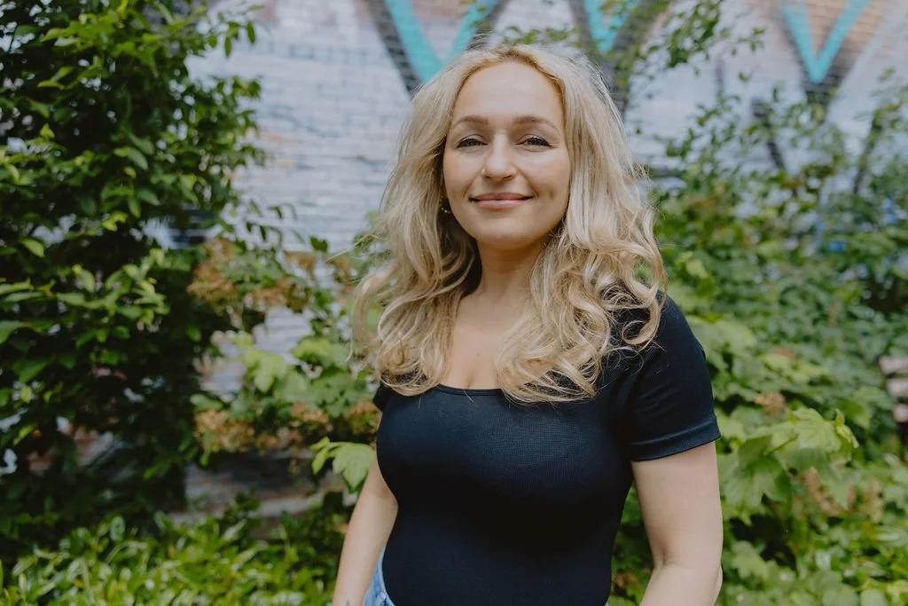 A headshot of Aneta. She is a woman with blonde hair wearing a black top poses confidently outdoors with greenery and a muralled brick wall behind her.