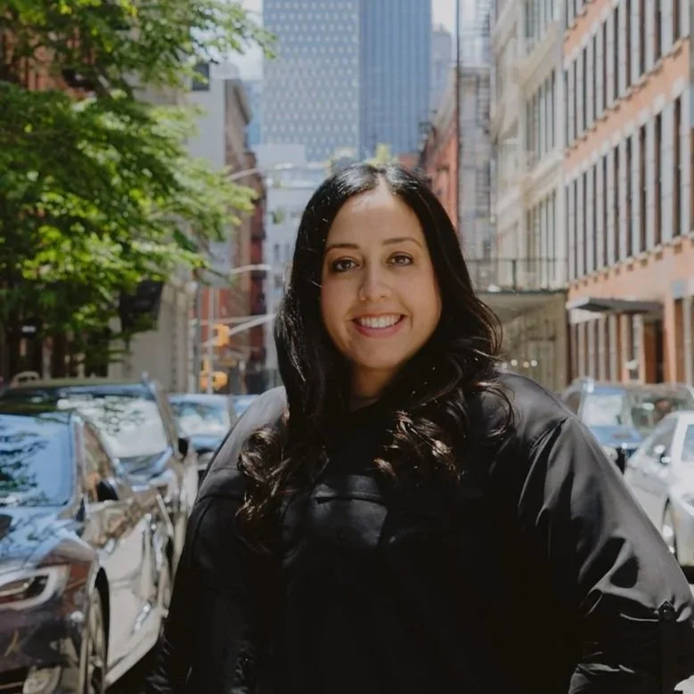 A headshot of Miriam. She is a woman with dark hair and is wearing a black shirt posing confidently on a cobbled New York City street lined with parked cars and the city skyline in the background.