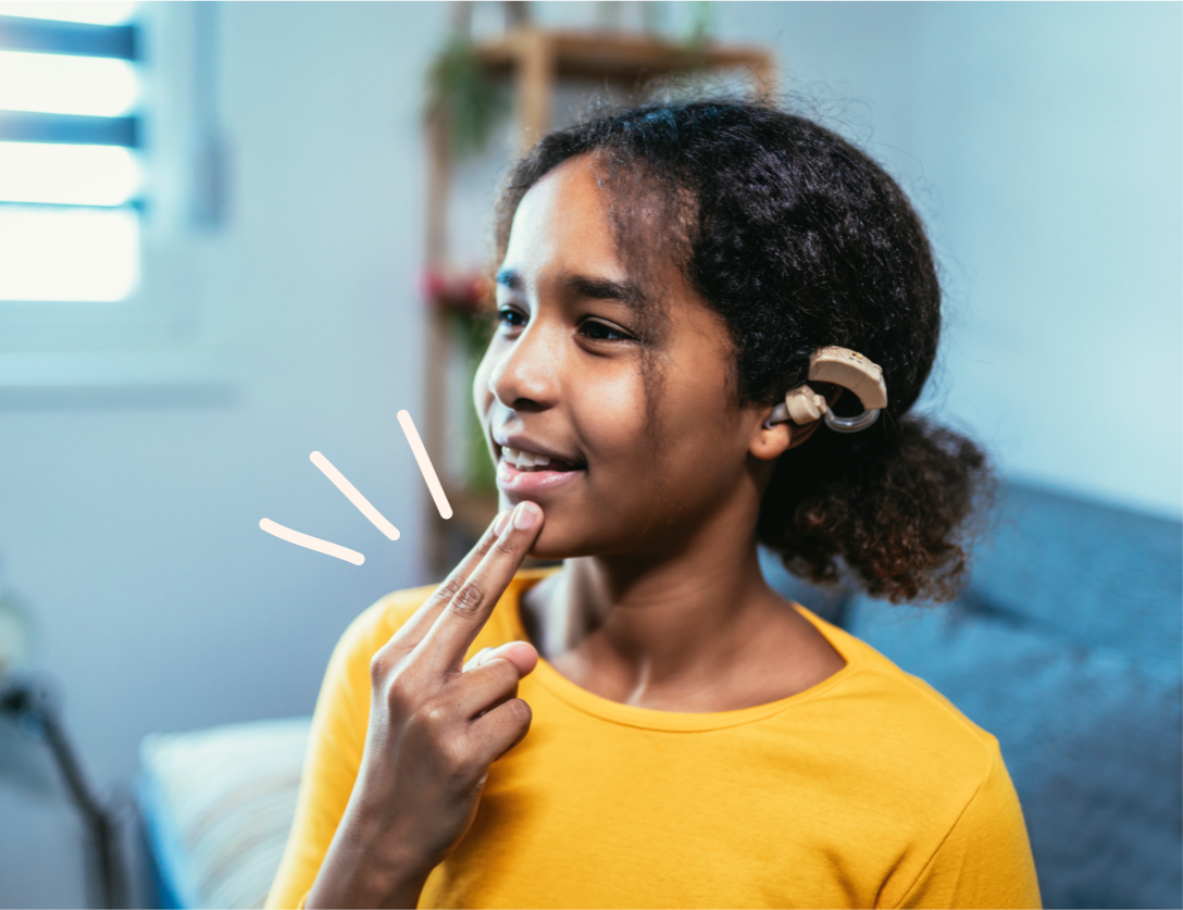 A girl wearing a cochlear implant and a yellow top uses two fingers to her lips as she signs, with playful doodle lines highlighting the motion. Soft indoor lighting and a cosy living space set the scene.