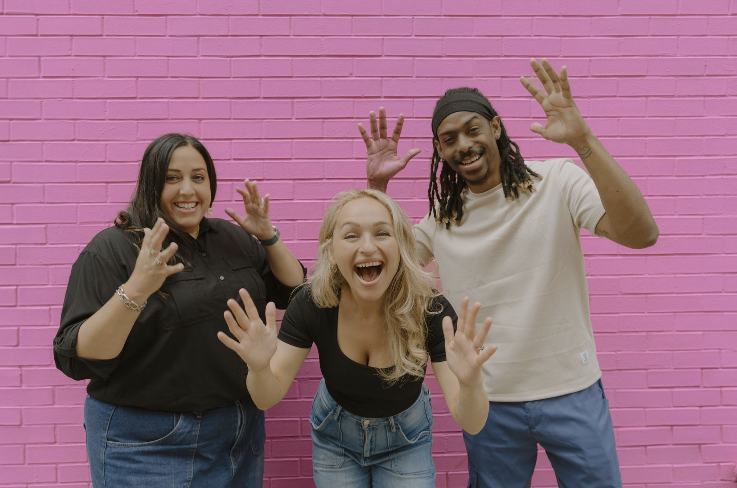 Miriam, Aneta, and Robert pose playfully with their hands up in friendly waves and signs against a vivid pink brick wall. They are a diverse group of young adults smiling in casual streetwear and joyful expressions conveying community and fun.
