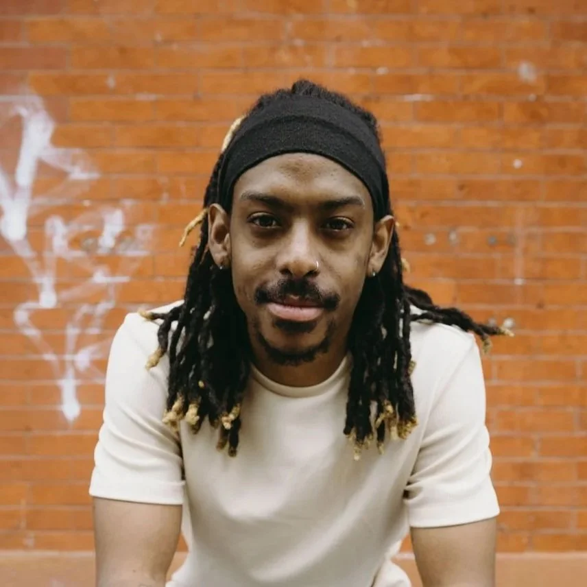 A headshot of Robert, he is in a cream tee shirt with an orange brick wall and street details forming an urban backdrop behind him.