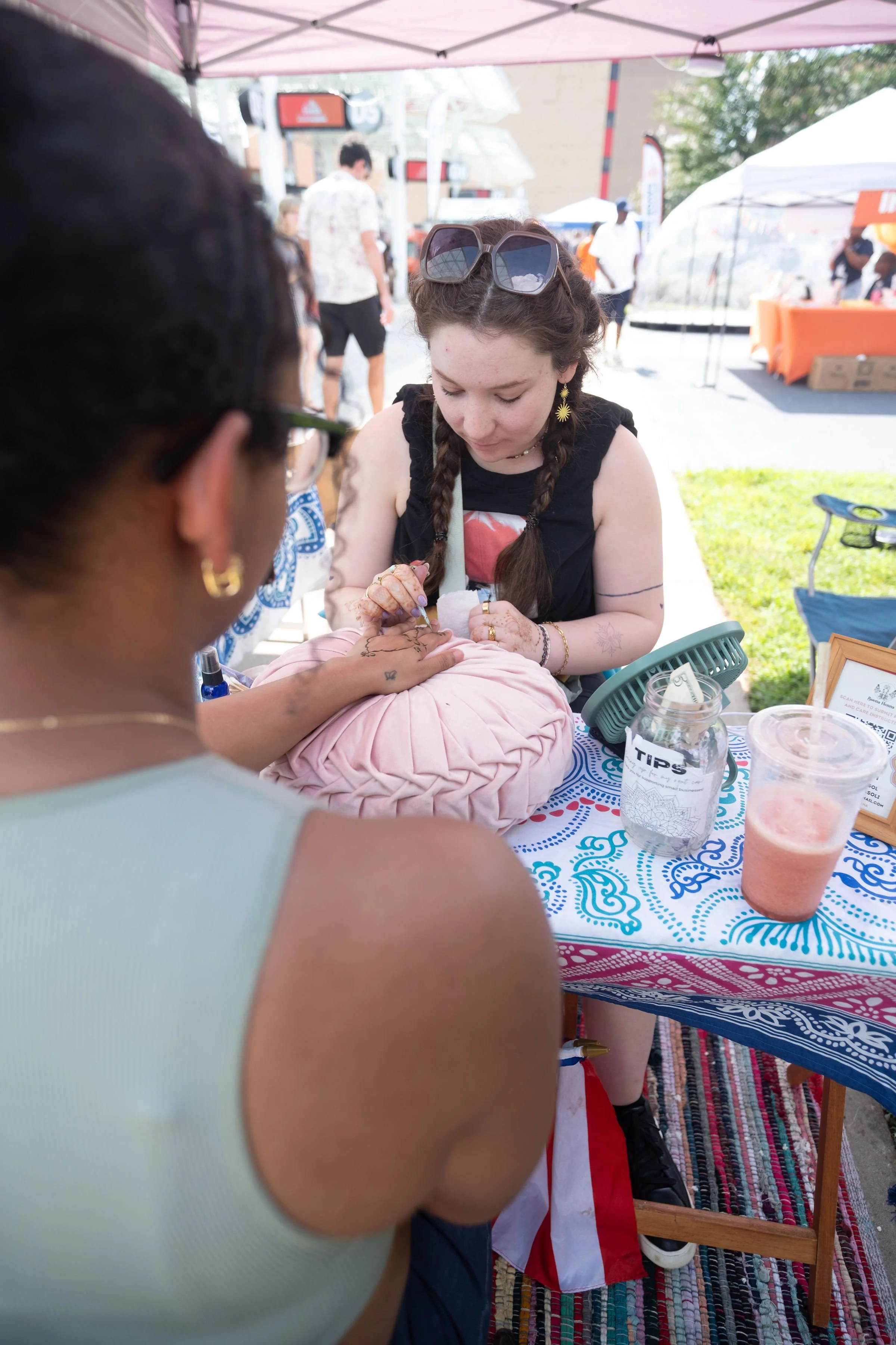 A person getting a tattoo on their hand at an outdoor event. There are tents, chairs, and other people in the background.