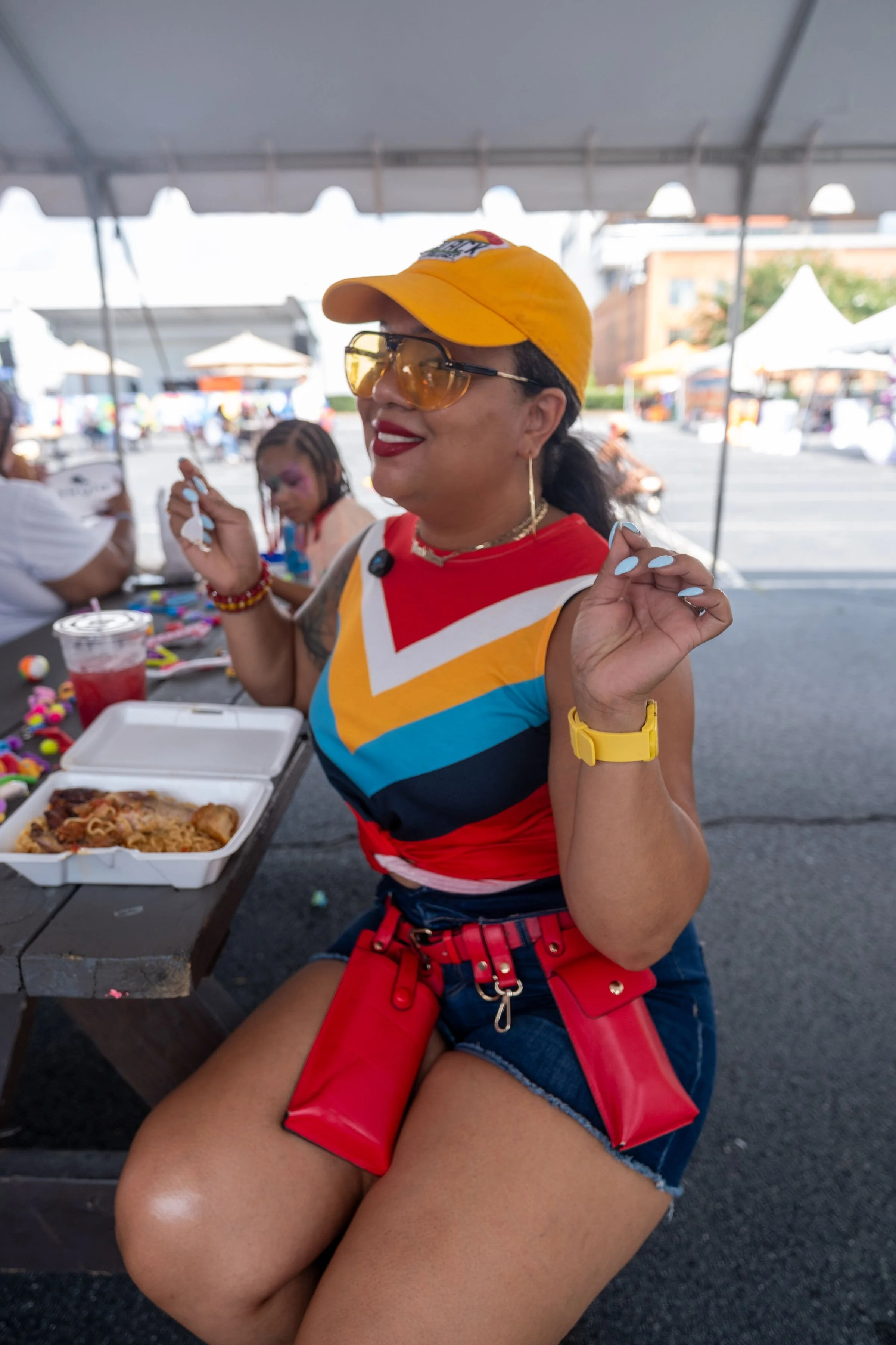 A woman smiling at a table with takeout food at an outdoor event. She is wearing a yellow cap, orange-tinted glasses, and a colorful sleeveless top. She has red lipstick, jewelry, and a yellow watch. The background shows other attendees and tents in a parking lot.