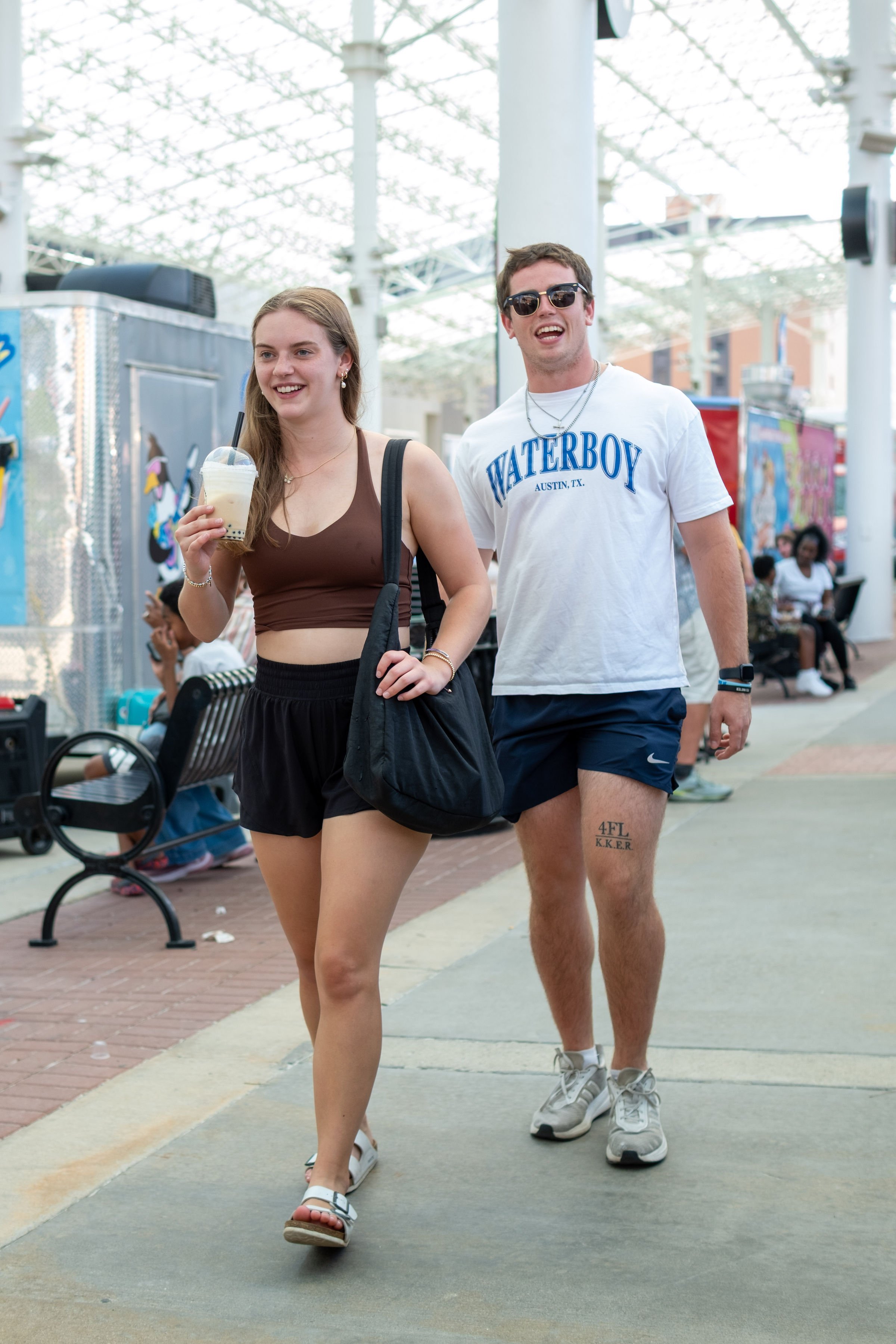 Two young people walking outdoors in a park or public space, smiling; the woman has long hair, is wearing a brown crop top, black shorts, and sandals, holding an iced beverage; the man has short hair, sunglasses, a white T-shirt with 'Waterboy Austin, TX', and athletic shorts, wearing sneakers.