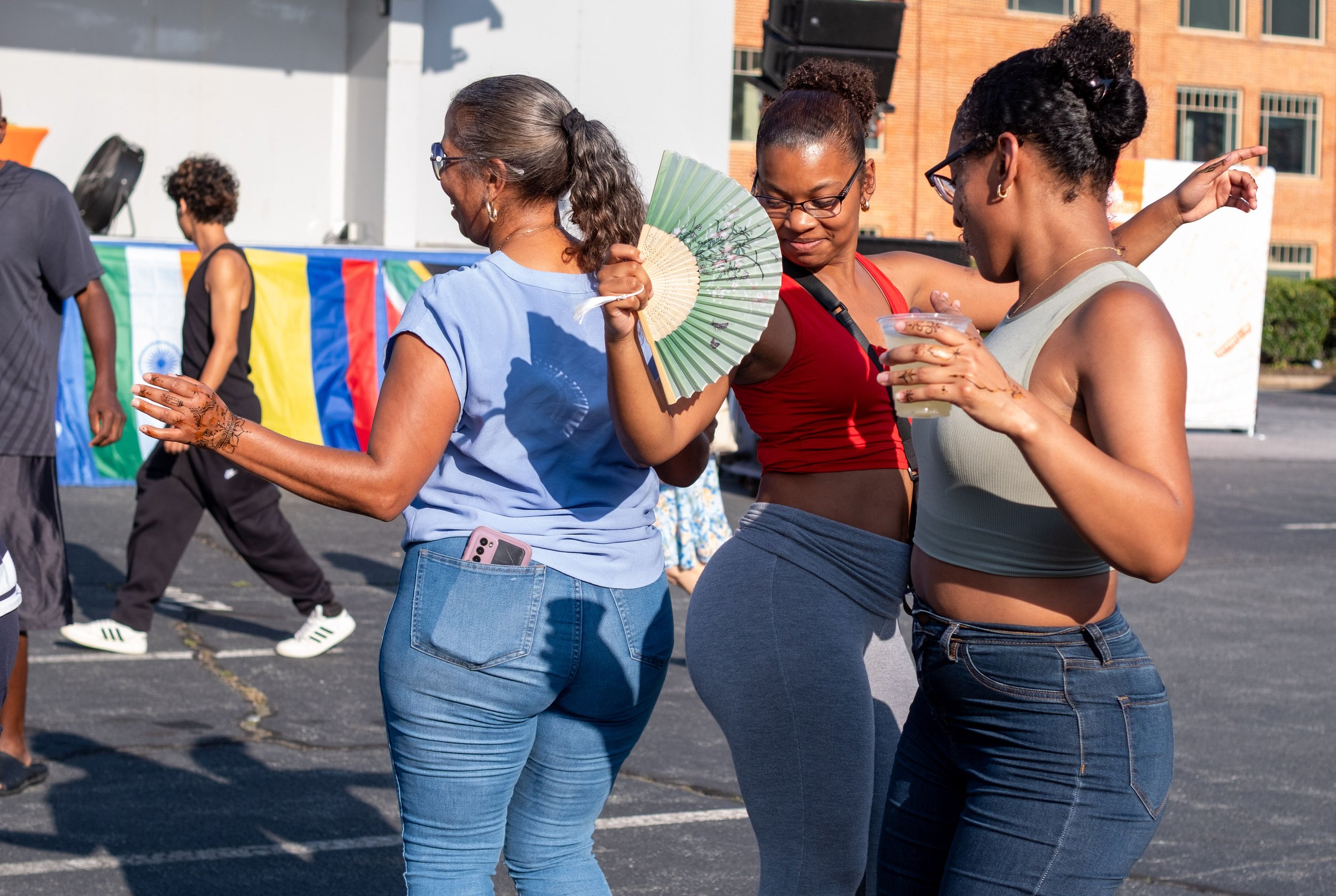 Three women dancing together outdoors at a festival, with a colorful flag display in the background.