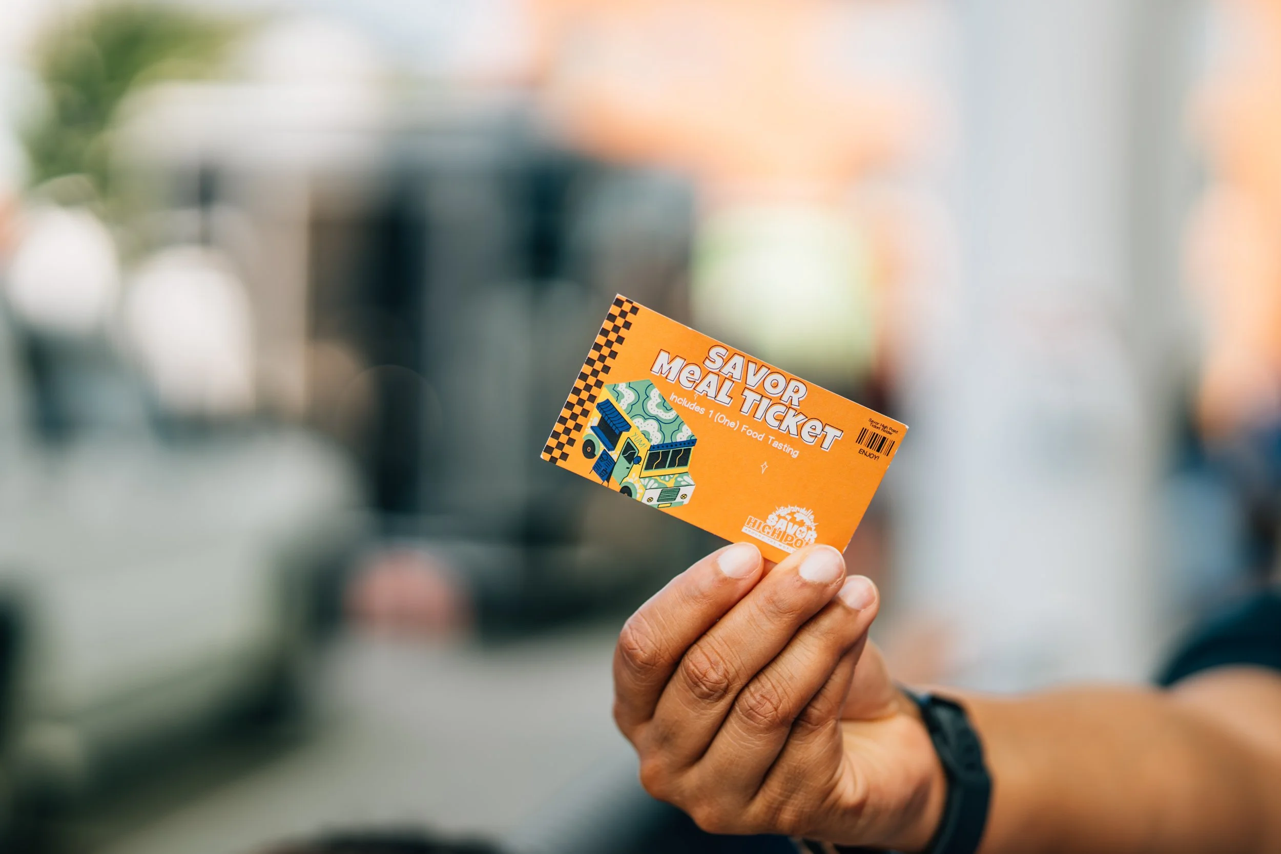 Person holding an orange 'Savor the Taste' food tasting ticket with a colorful design, in a blurred indoor setting.