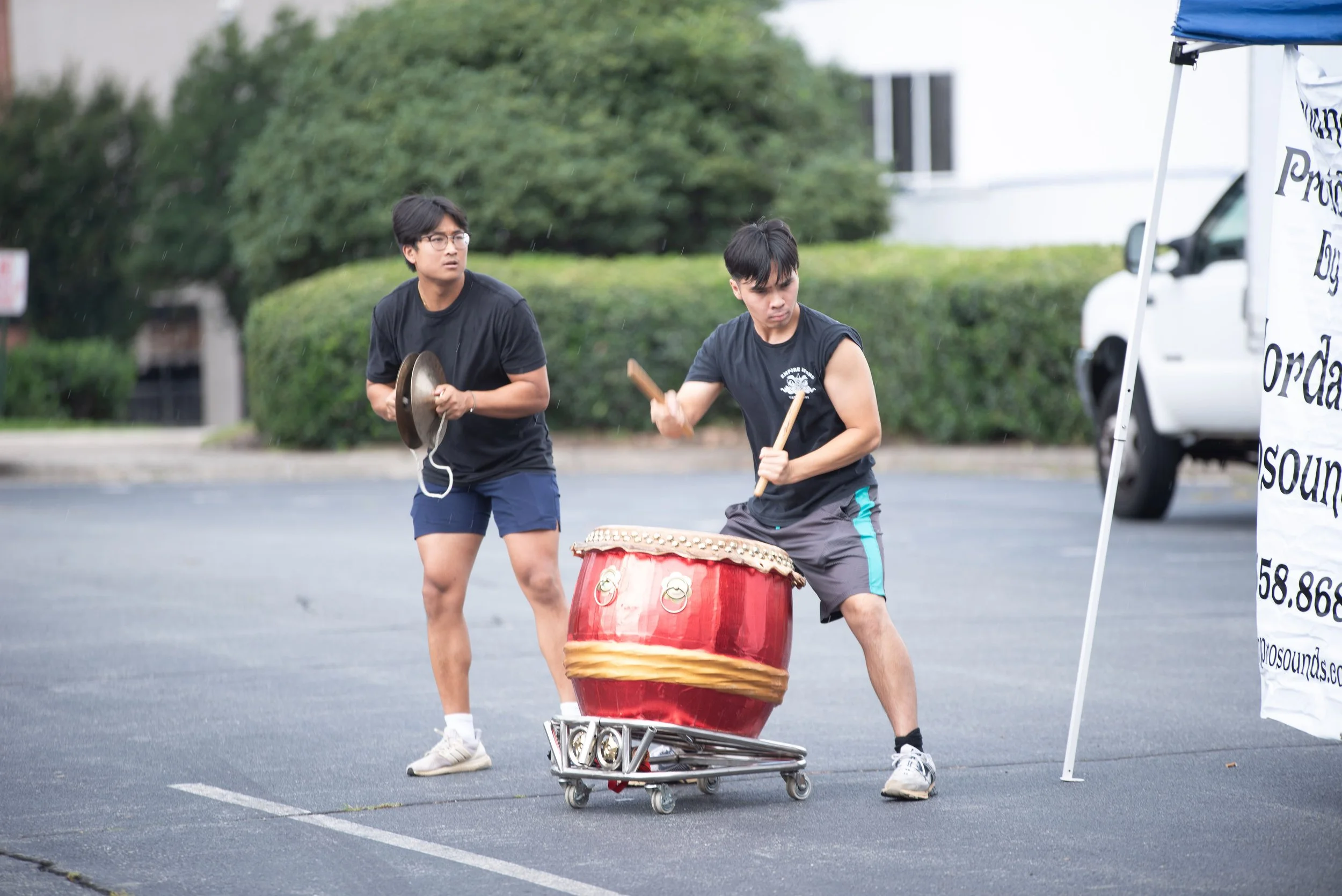 Two young men performing at an outdoor event, playing a drum and cymbals, with a large traditional drum on a wheeled stand, in a parking lot during rainy weather.
