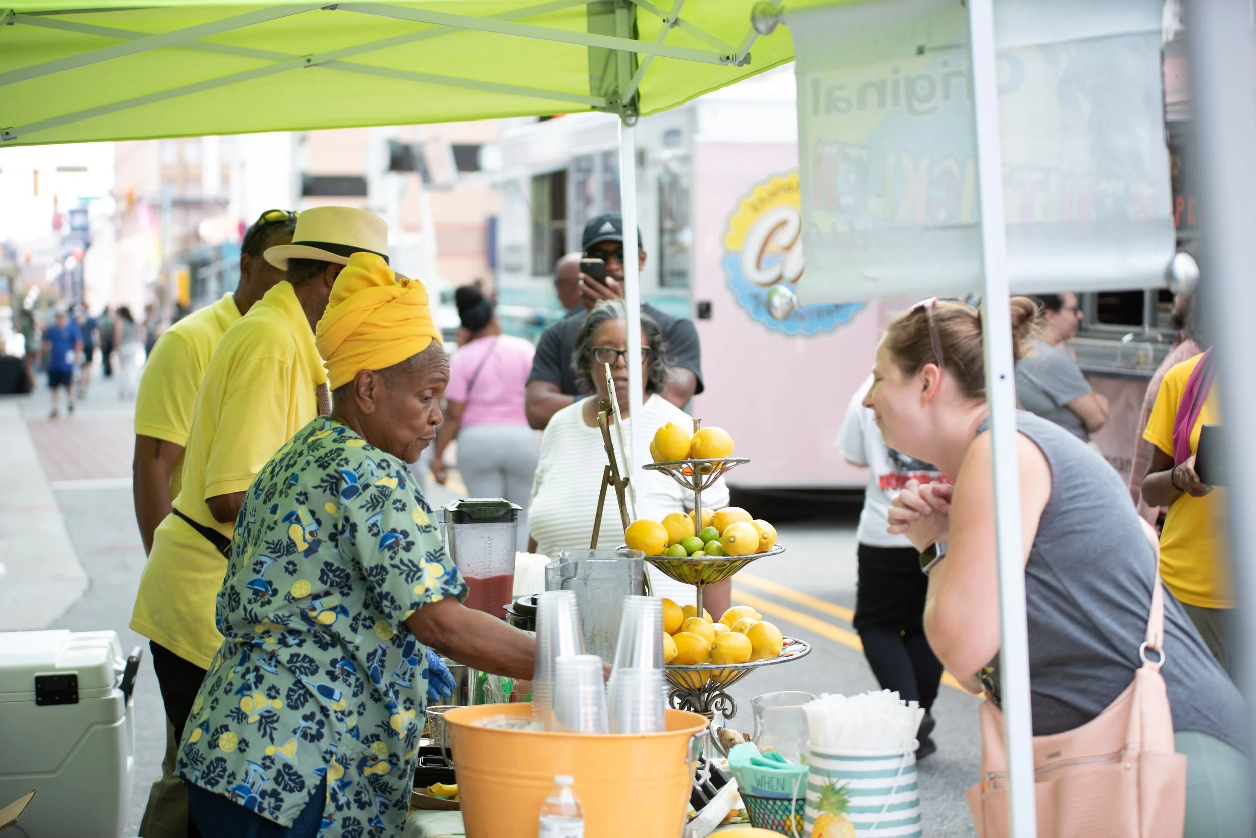 People at a fruit stand on a city street with a green canopy, selling citrus fruits such as lemons and limes