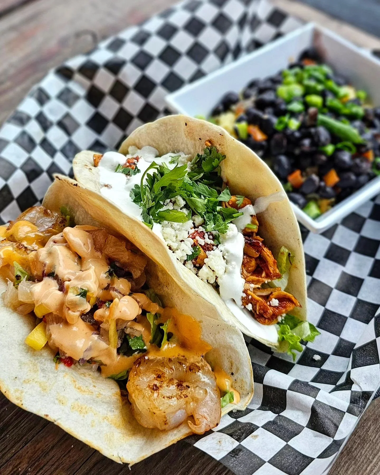 Two tacos filled with shredded meat, vegetables, cheese, and sauce, on black-and-white checkered paper.