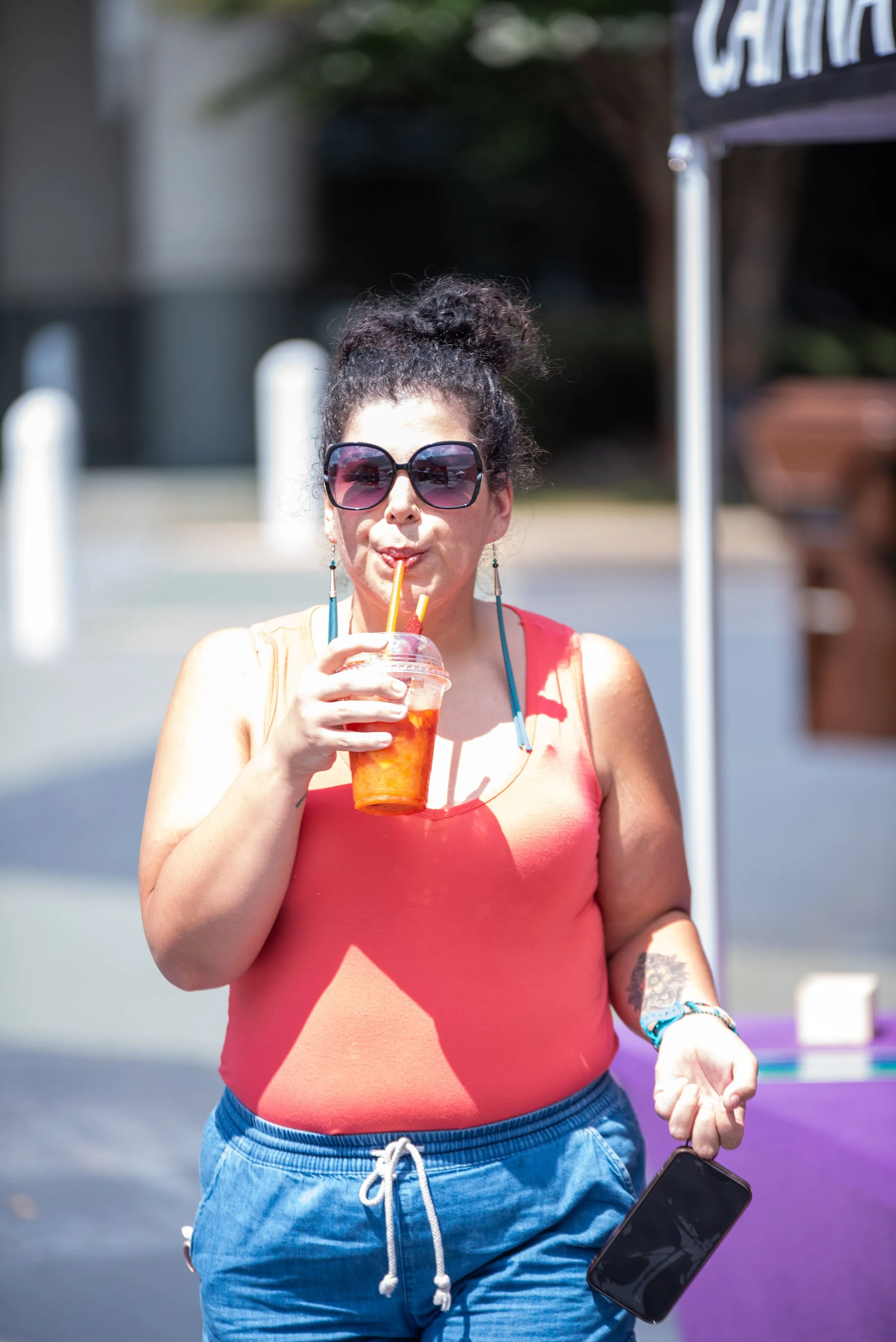 Woman with curly hair in a bun wearing sunglasses, a sleeveless orange top, and blue shorts, sipping a drink through a straw outdoors.
