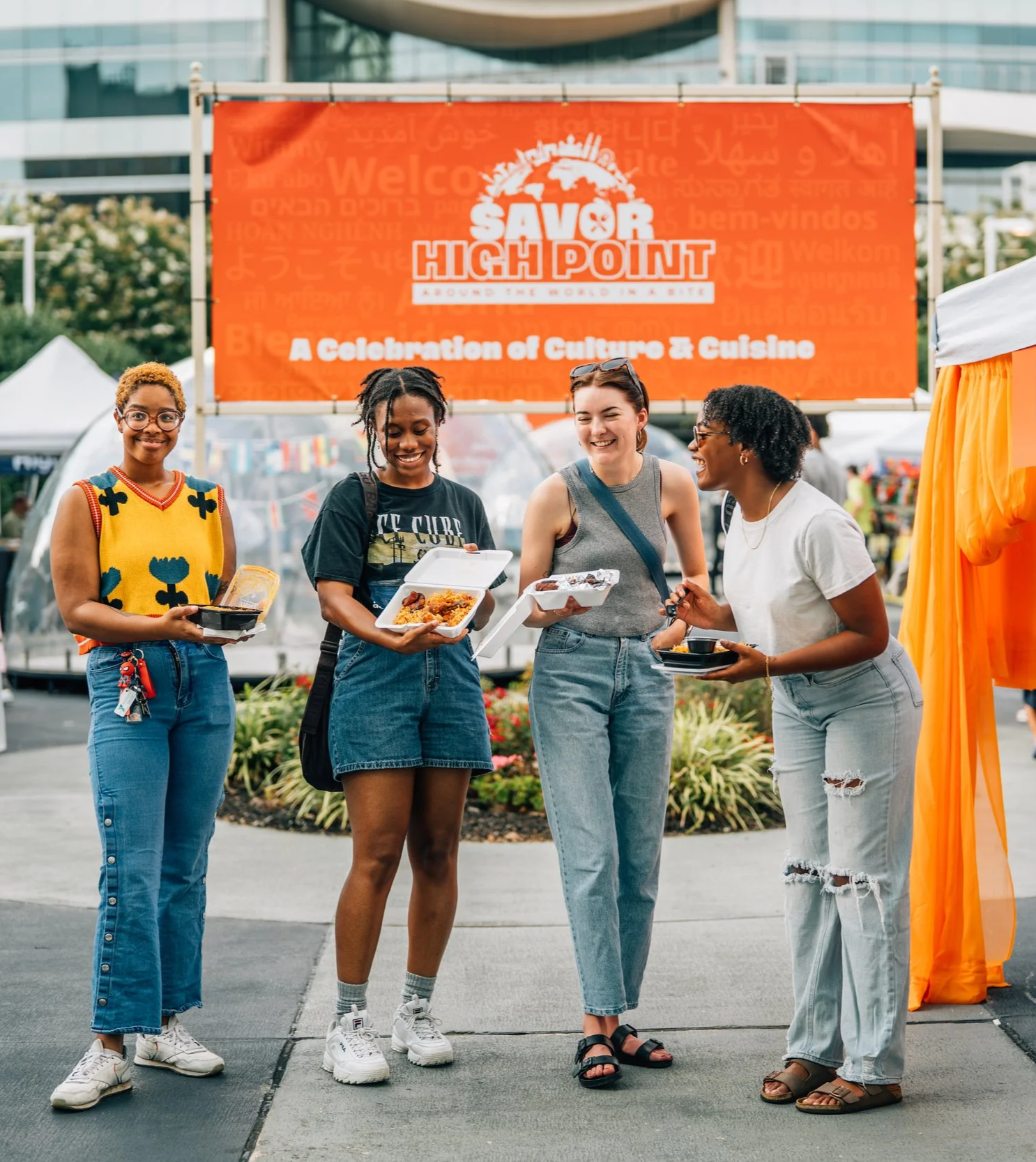 Four women standing outdoors at a food festival under an orange 'Savor High Point' banner. They are smiling, holding food containers, and enjoying the event.