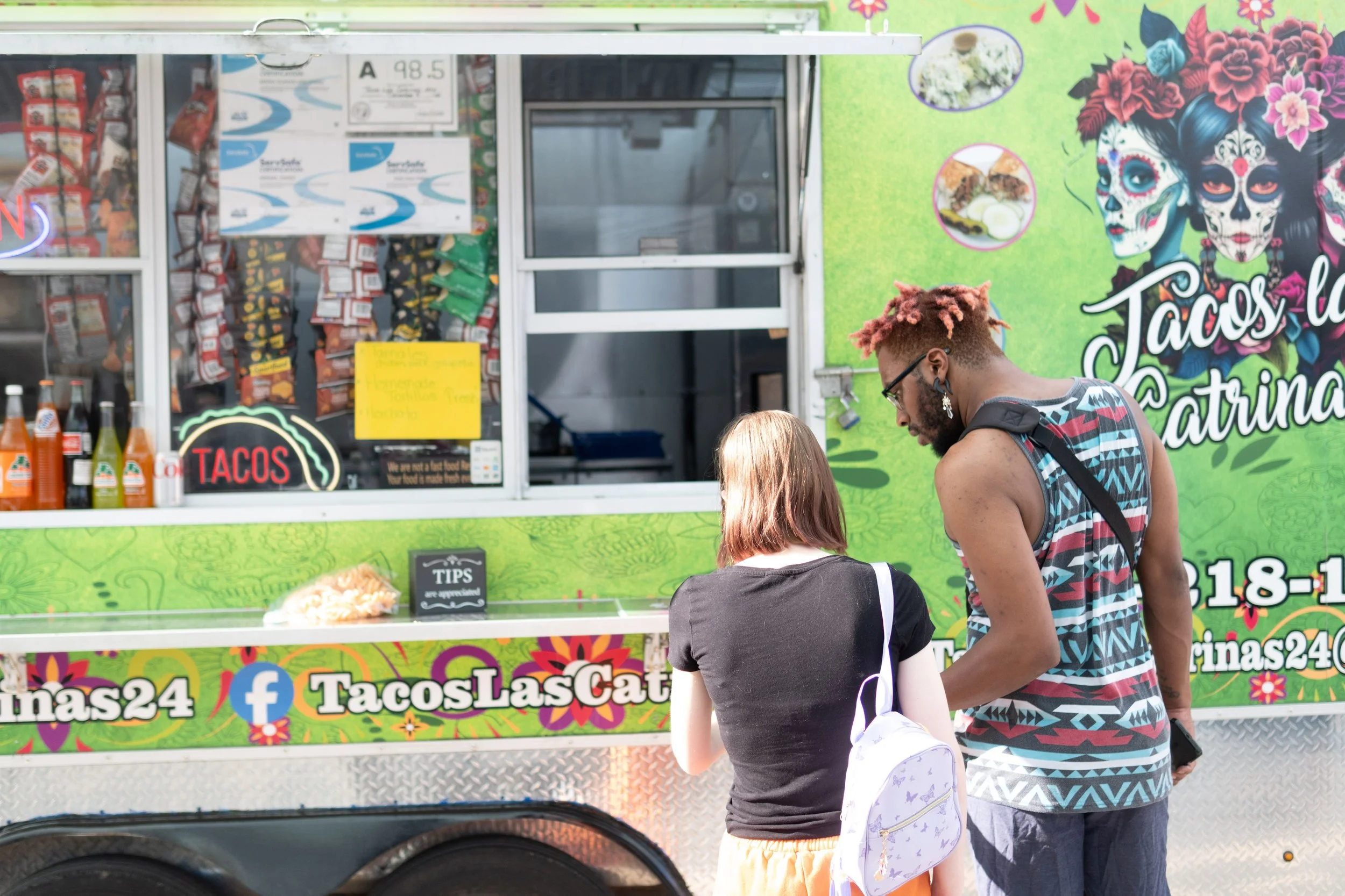 A colorful food truck with green, pink, and black design featuring Day of the Dead imagery and the words "Tacos" and "Tacos Las" displayed. People, including a man with pink-dyed hair and a woman with brown hair and a young girl with a backpack, are standing in front of the truck.