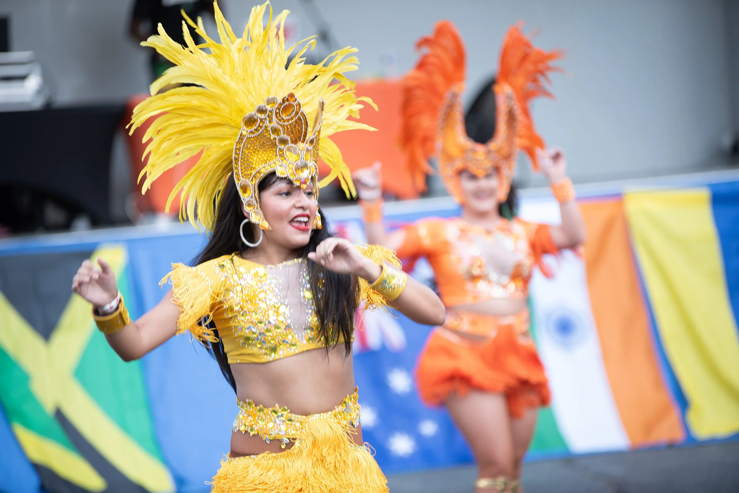 Two women dancing in colorful costumes during a cultural event. The woman in the foreground wears a yellow feathered headdress and midriff top, while the woman in the background wears an orange costume with a feathered headpiece.